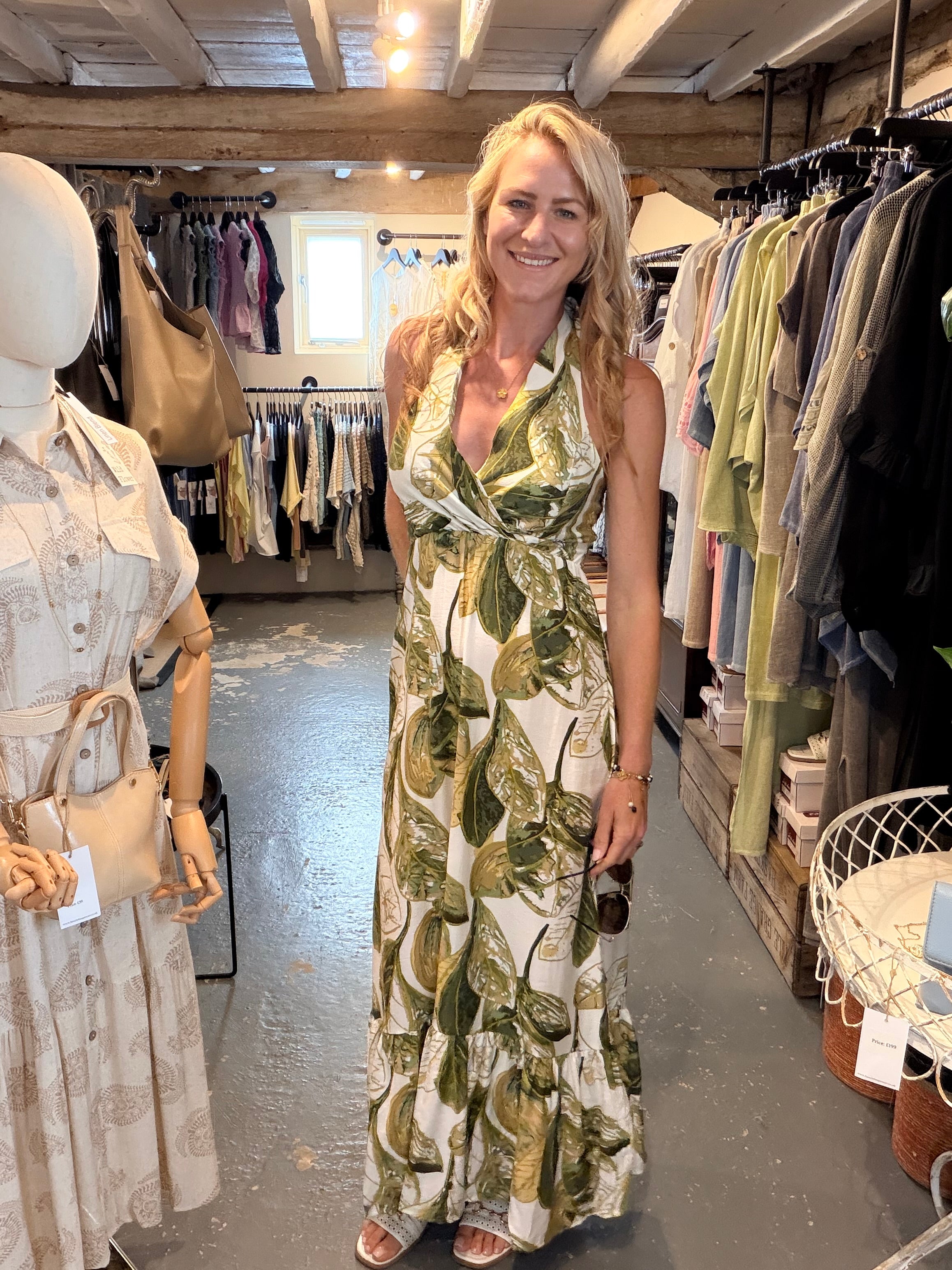 A woman stands in a boutique wearing a sleeveless halter maxi dress with a green leaf print on a cream background. The shop has clothing racks, mannequins, and a rustic wooden ceiling.