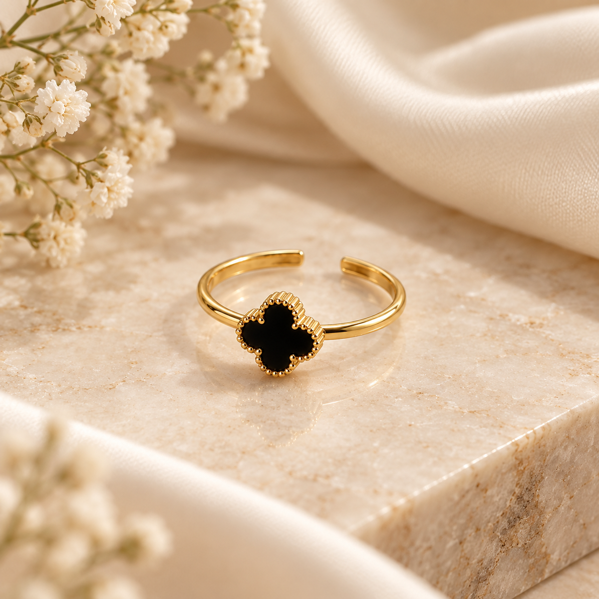 A gold-tone adjustable ring with a black clover-shaped stone, styled on a beige marble block with white fabric and baby's breath flowers.