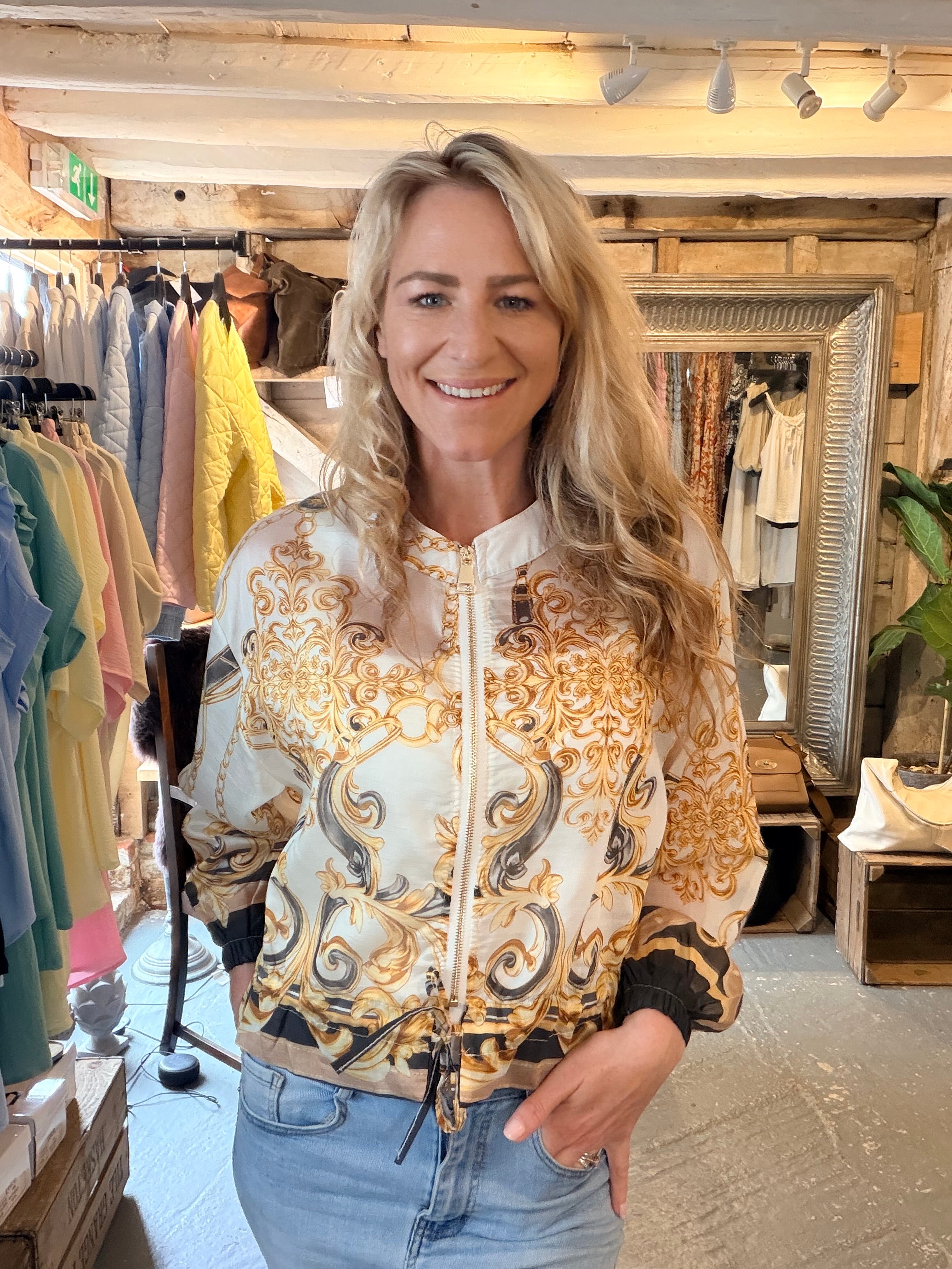 A woman with blonde hair smiling in a boutique, wearing a cream-colored bomber jacket with an ornate gold and black baroque pattern, paired with light blue jeans.