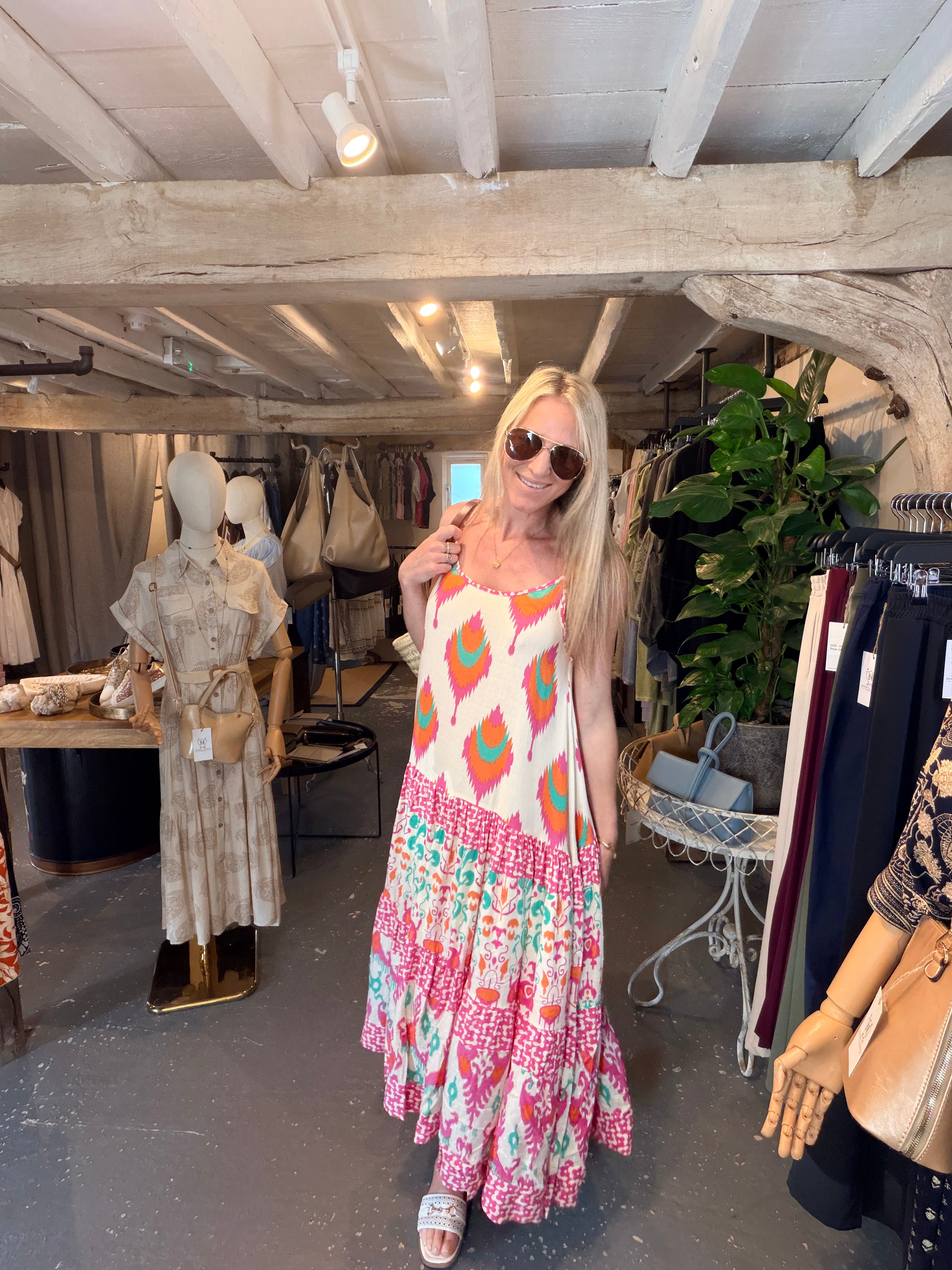 A woman wearing a colorful bohemian-style tiered maxi dress with geometric and floral patterns, standing inside a rustic clothing boutique.