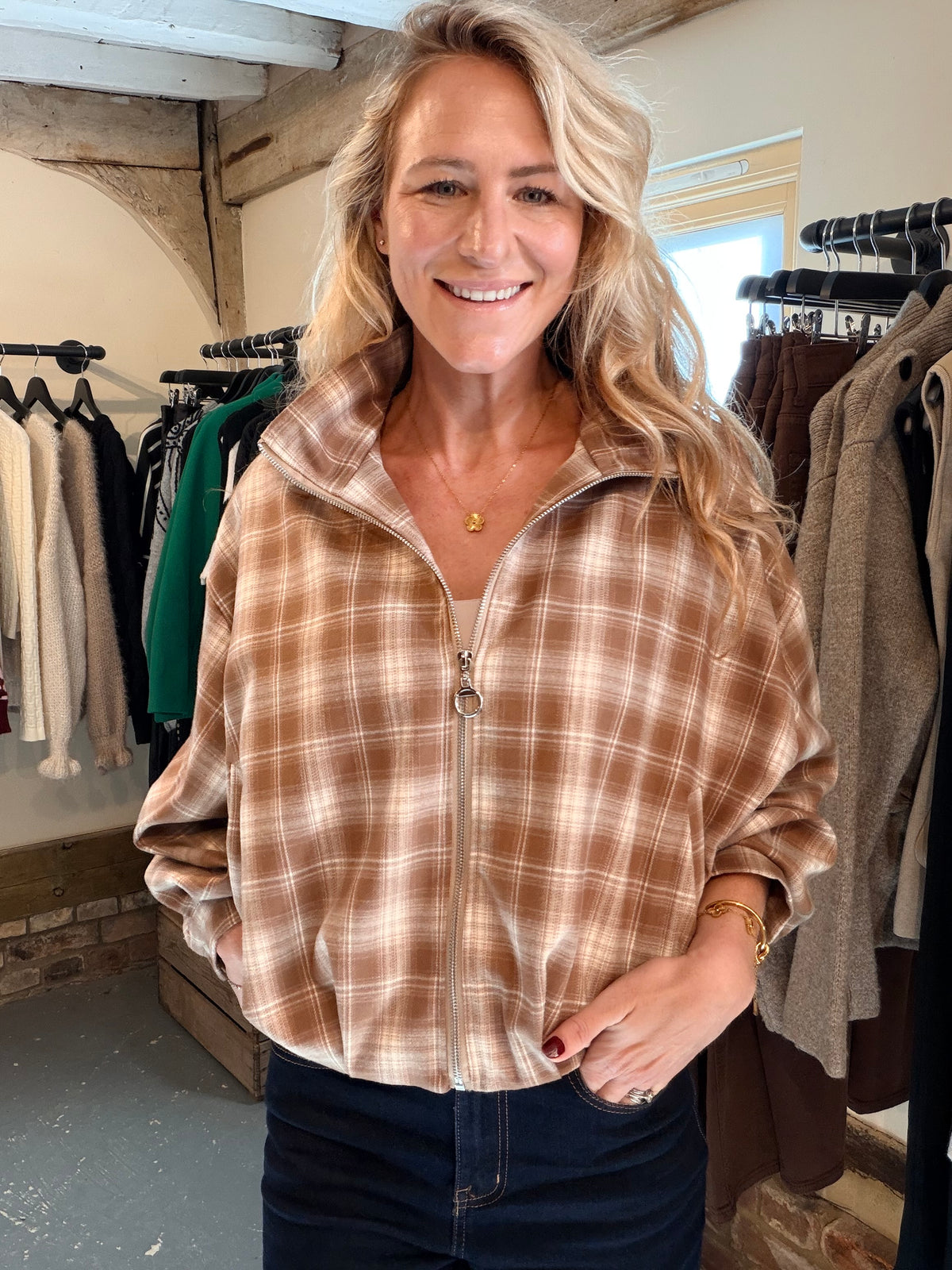 A smiling blonde woman wearing a brown and beige plaid zip-up jacket with dark blue jeans, standing inside a clothing boutique with racks of garments in the background.