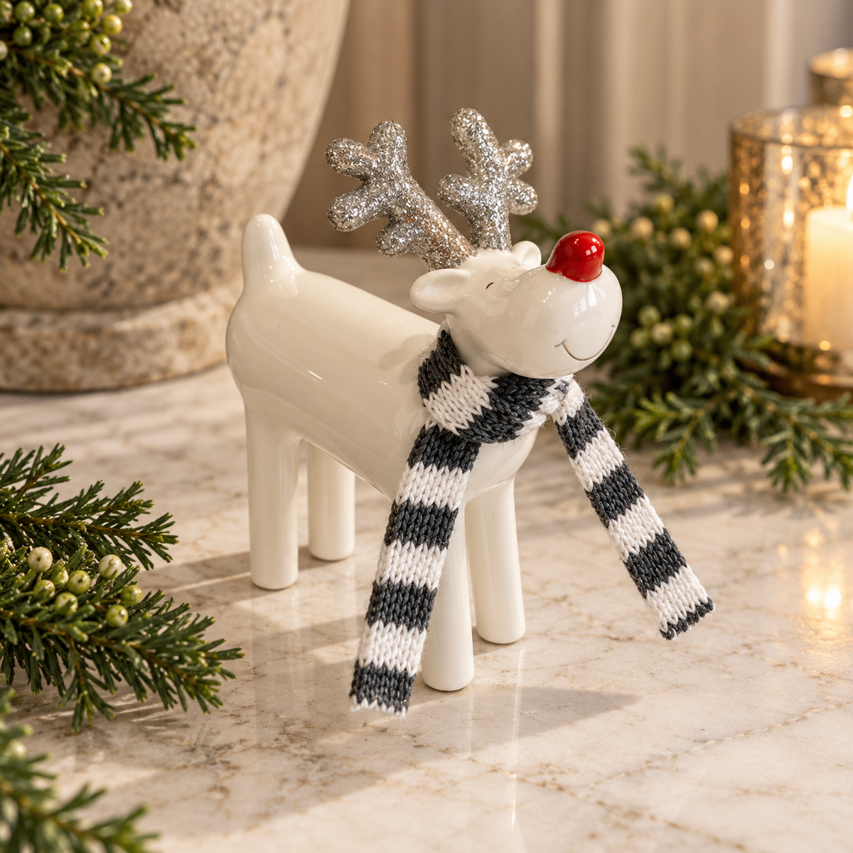 A white ceramic Christmas reindeer figurine with silver glitter antlers, a black and white striped knit scarf, and a red nose, displayed on a marble surface with holiday greenery and a candle in the background.