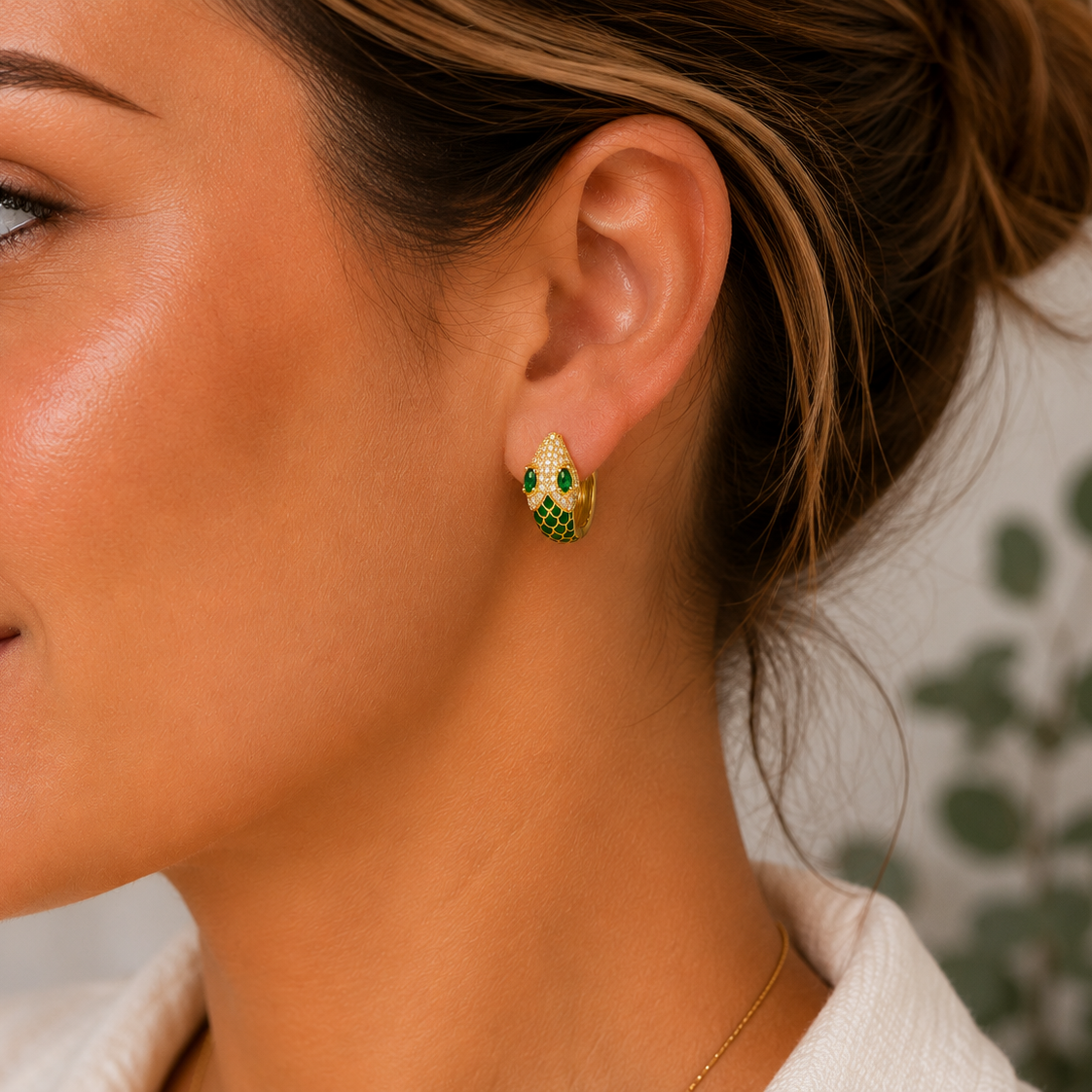 A close-up lifestyle shot of a woman wearing a small gold huggie hoop earring adorned with green enamel and tiny clear stones.