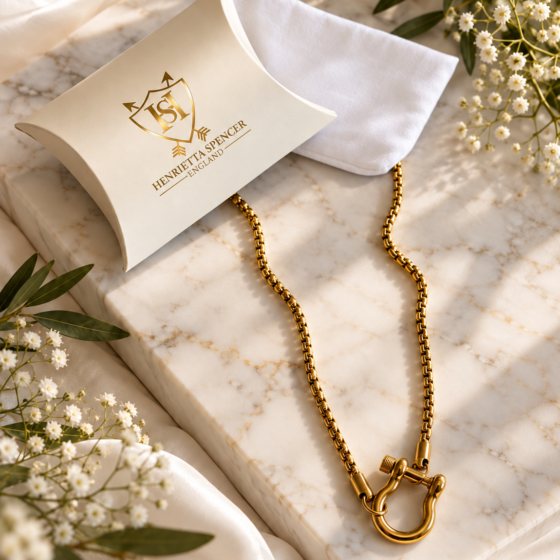 A gold-tone chain necklace with a large ring clasp is displayed on a white marble slab with a branded cream-colored presentation pouch and a white microfiber cloth. The scene is styled with white baby's breath flowers and green leaves on draped white fabric.