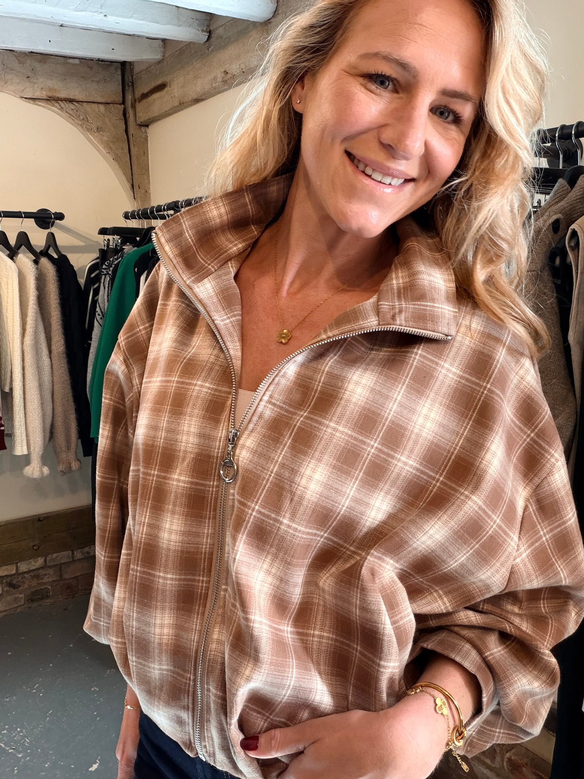 A smiling blonde woman wearing a brown and beige plaid zip-up jacket in a boutique setting.
