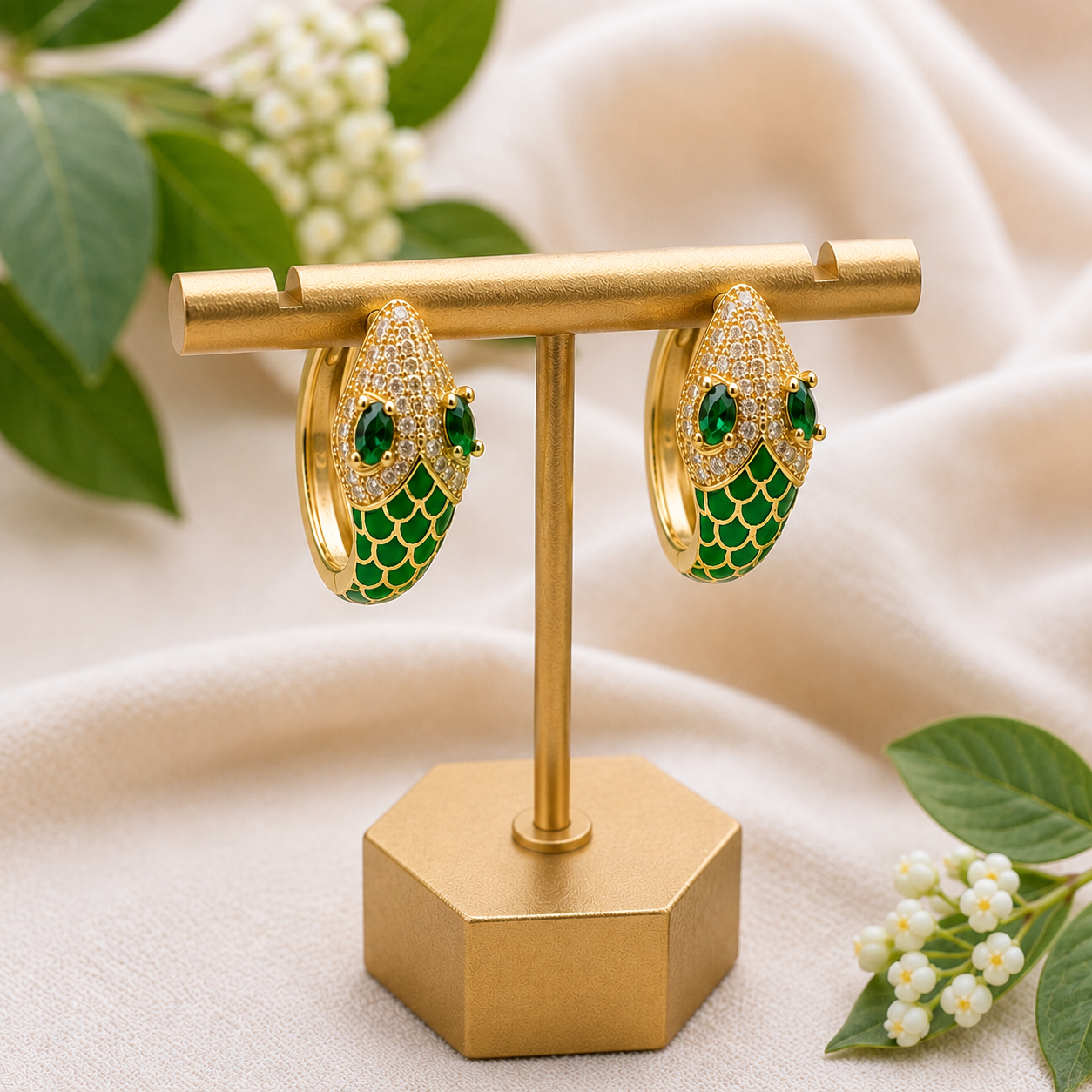 A pair of gold hoop earrings with green enamel scales and rhinestone details, displayed on a gold stand against a soft beige fabric backdrop with green leaves and small white flowers.