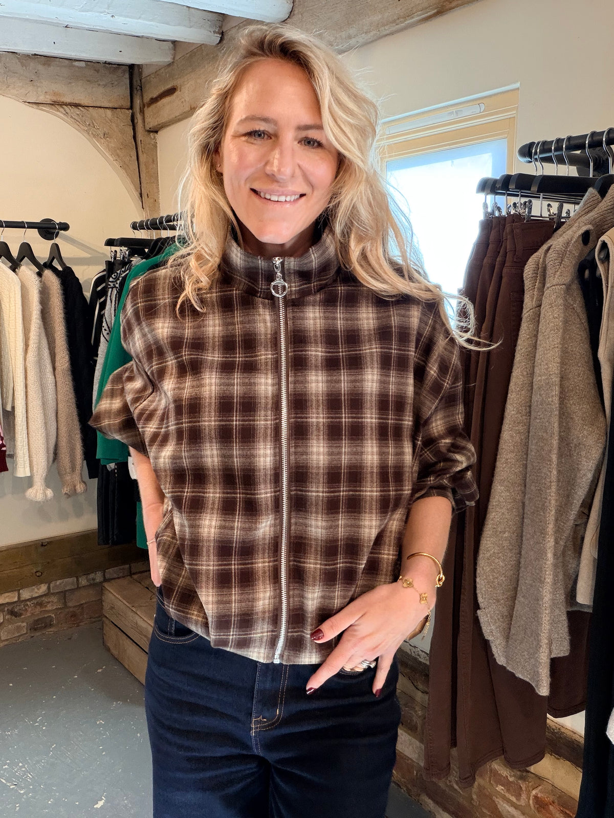 A smiling blonde woman wearing a brown and beige plaid zip-up fleece jacket with dark blue jeans, standing inside a clothing boutique with racks of garments in the background.
