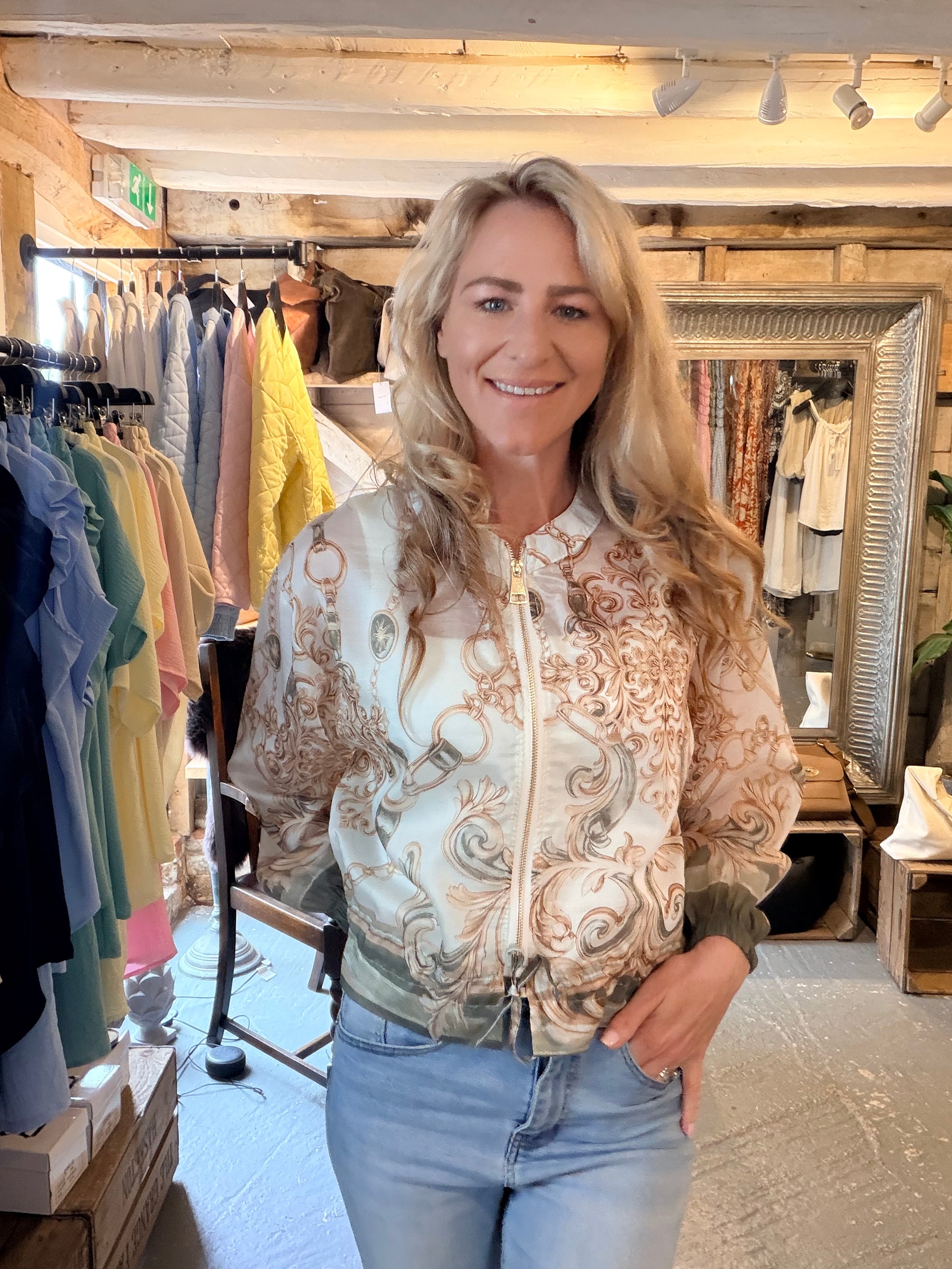 A smiling blonde woman wearing a cream and brown damask-patterned zip-up jacket and light blue jeans, standing inside a clothing boutique.