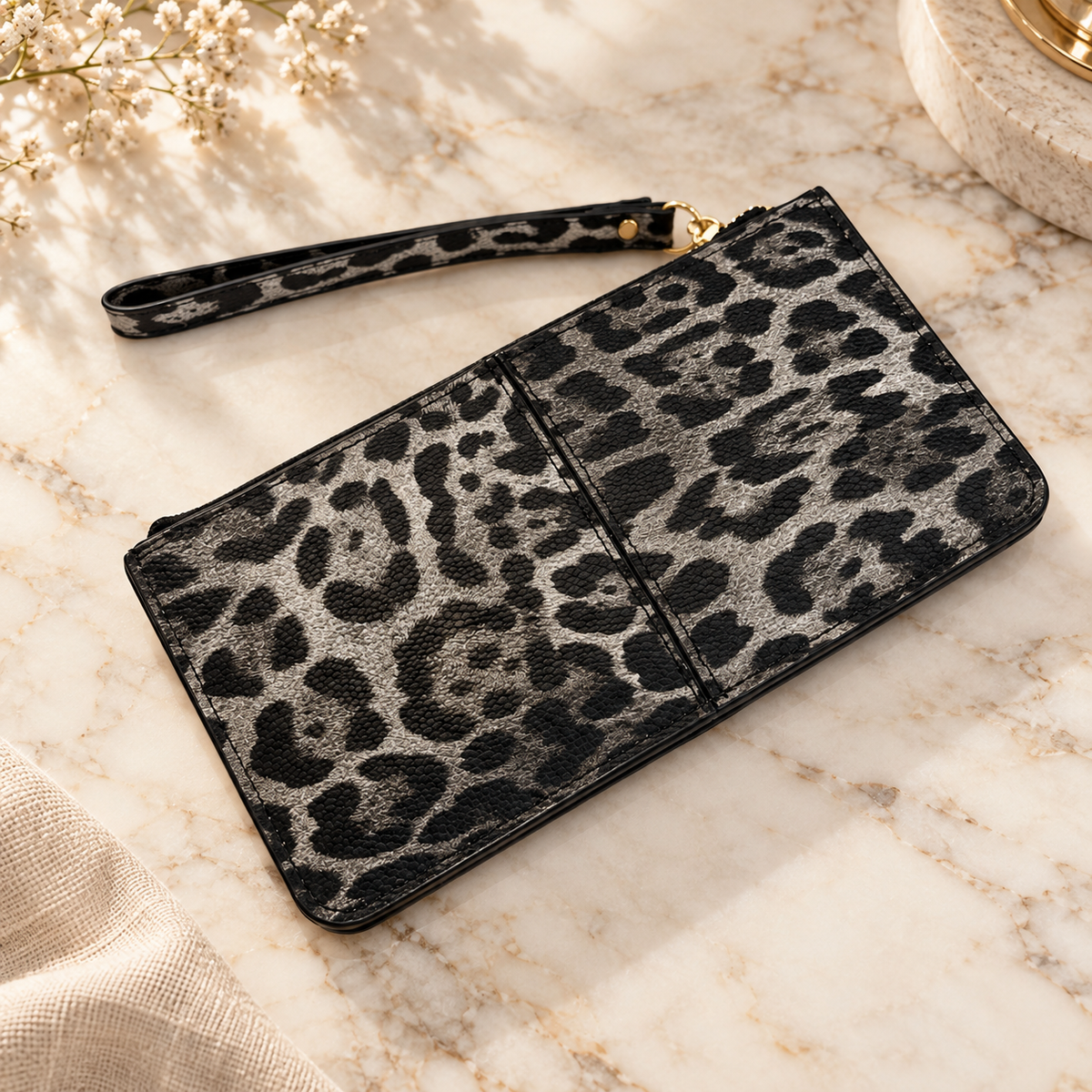 A leopard print wristlet clutch with a matching wrist strap, shown on a light marble surface with neutral decorative props.