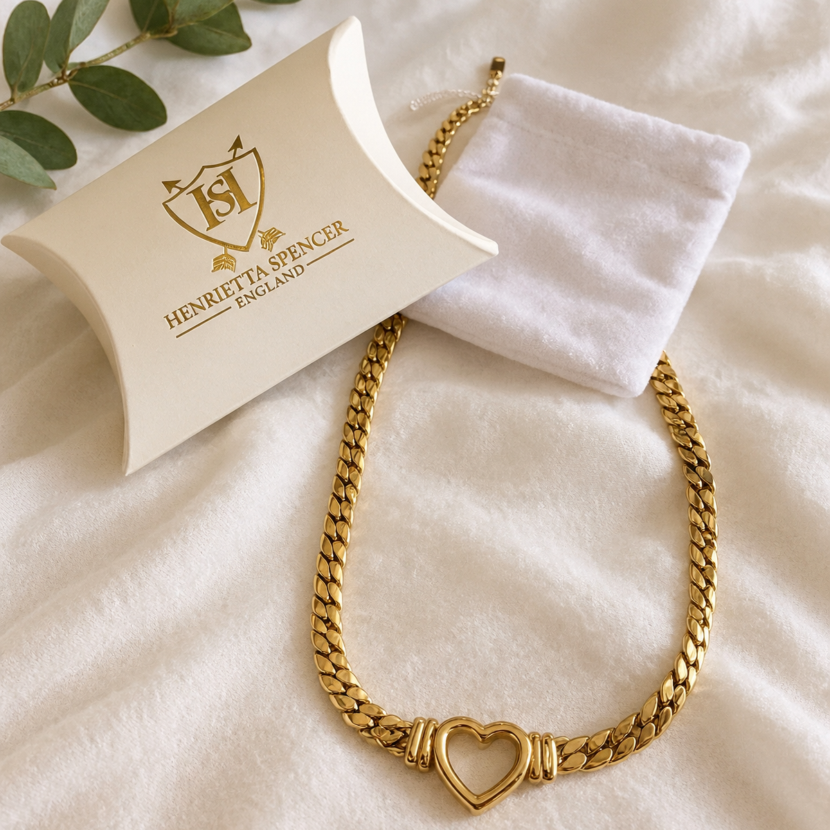 A gold heart-link chain necklace displayed on a soft cream fabric surface, with a white jewelry pouch and branded cream pillow insert. The brand name Henrietta Spencer England is visible on the insert.