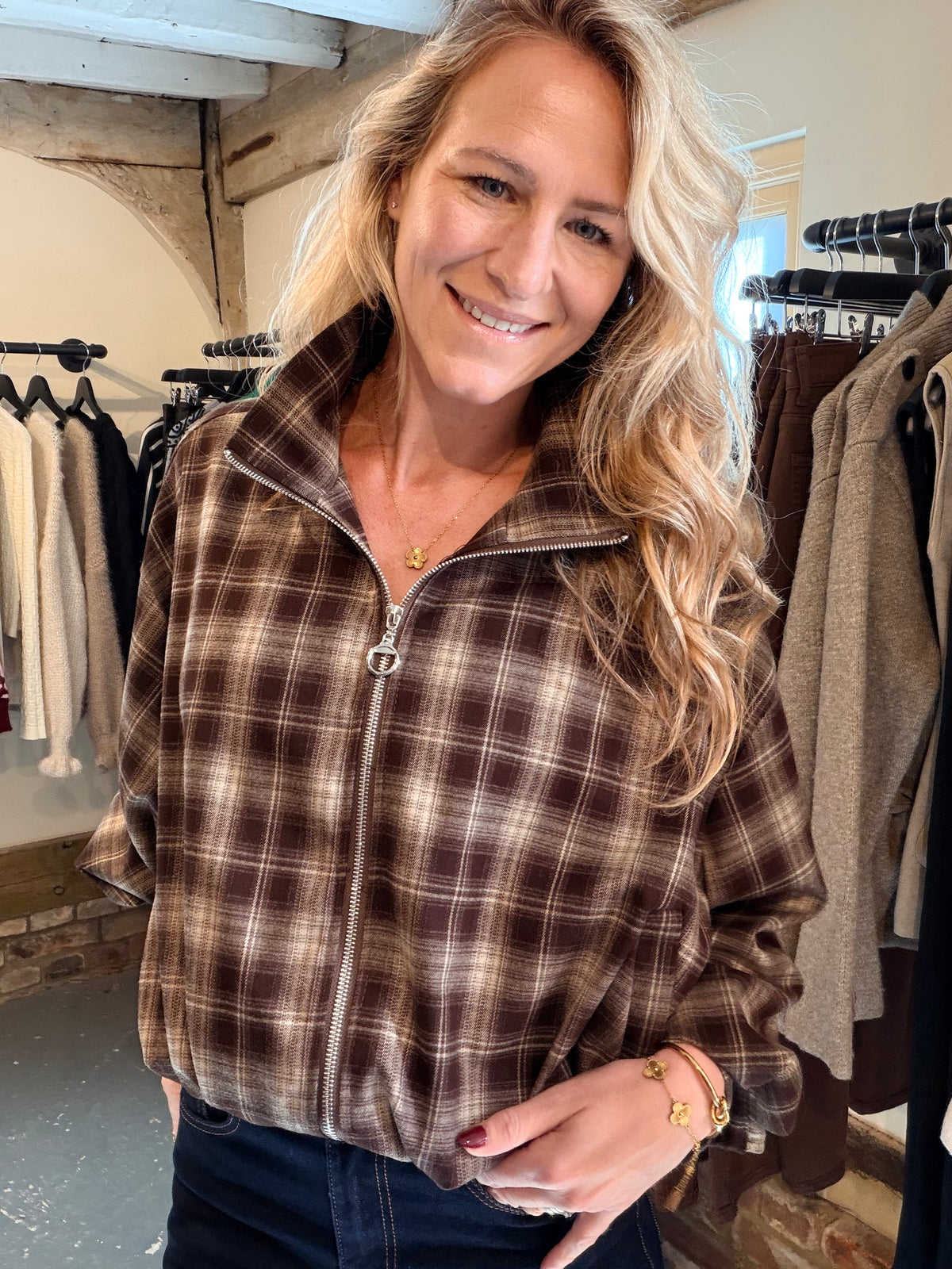 A smiling blonde woman wearing a brown and beige plaid zip-up fleece jacket with a stand collar, standing inside a clothing boutique with racks of garments in the background.