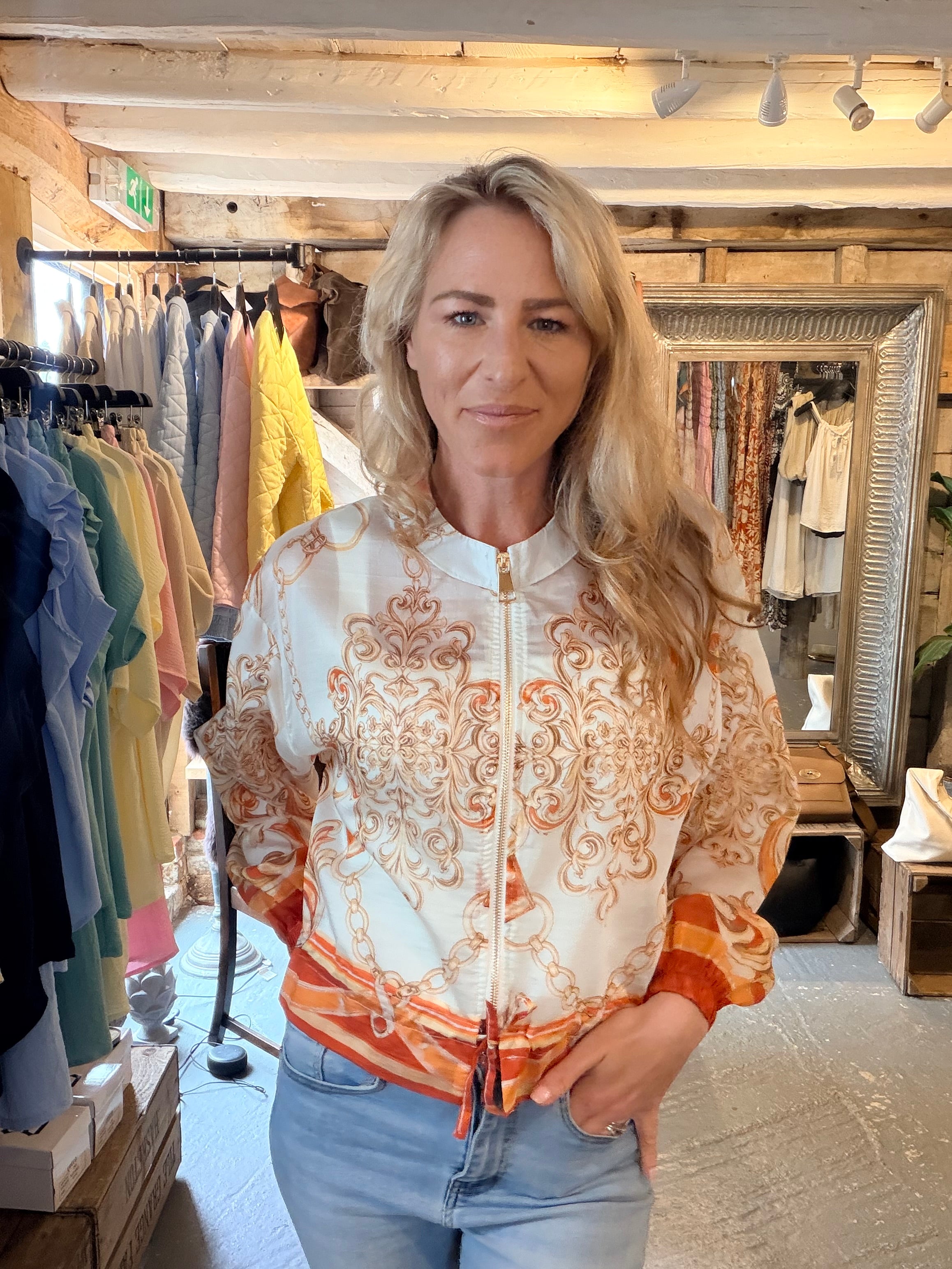 A woman with blonde hair wearing a white zip-up bomber jacket with an orange and brown baroque pattern, standing inside a clothing boutique.