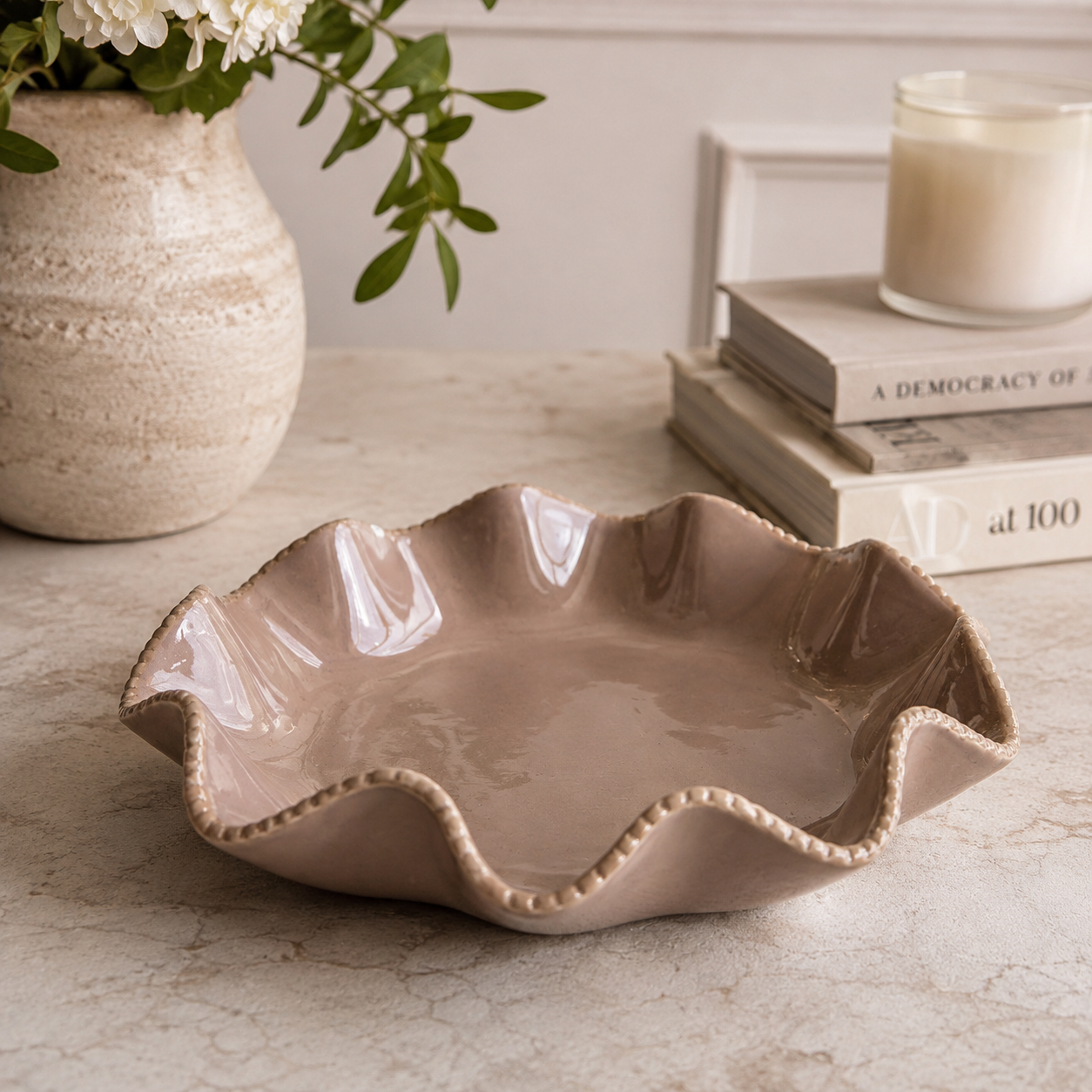 A scalloped ceramic bowl in a warm taupe-beige color, styled on a marble tabletop with a textured vase of flowers and books in the background.