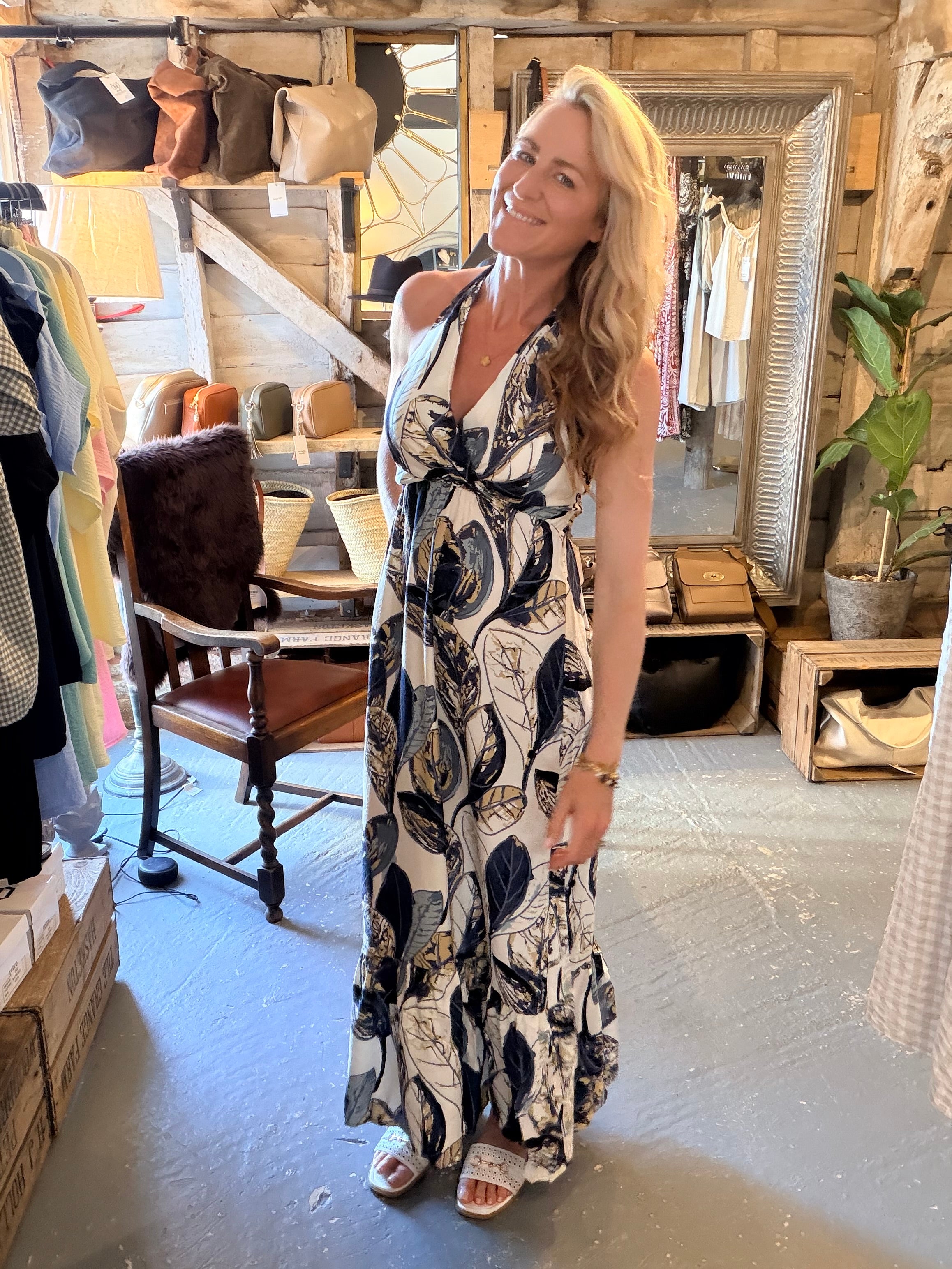 A woman wearing a sleeveless, V-neck maxi dress with a navy and beige botanical leaf pattern, standing inside a rustic clothing boutique.