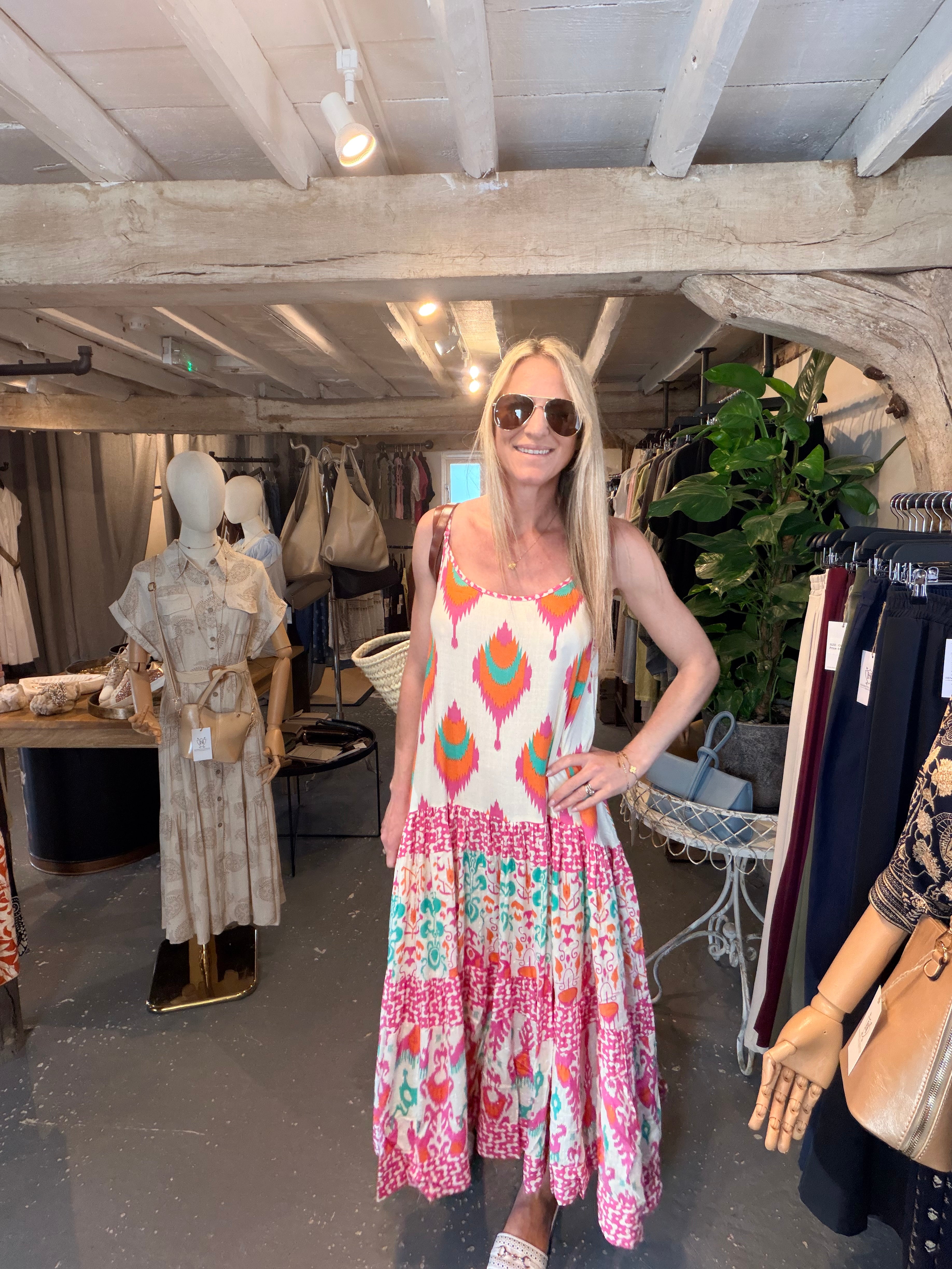 A woman wearing a sleeveless bohemian-style dress with a cream top and a pink, orange, and teal ikat patterned skirt, standing inside a boutique.