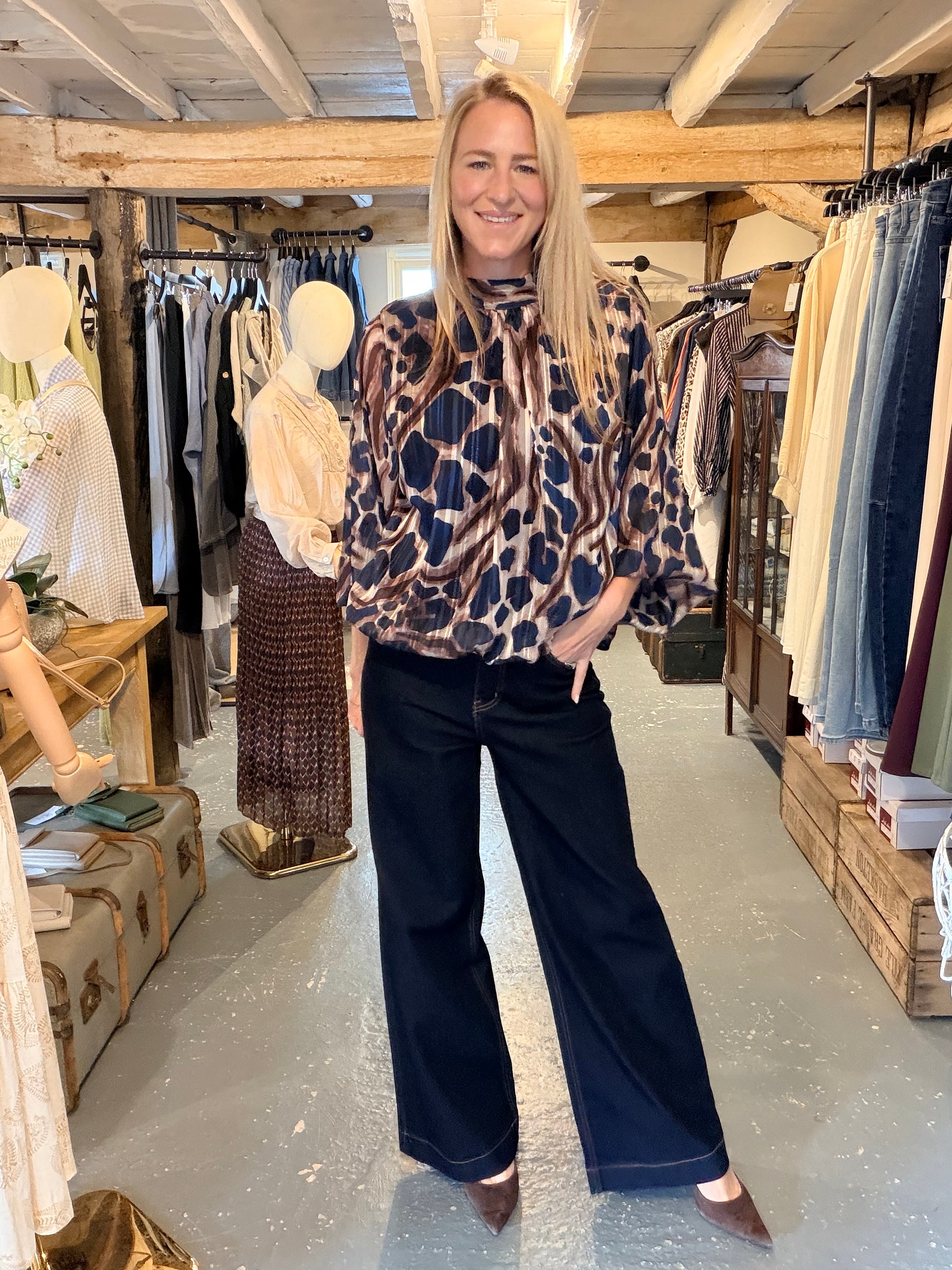 A woman stands in a clothing boutique wearing a brown, beige, and navy leopard-print blouse with wide-leg navy pants and brown pointed-toe flats.