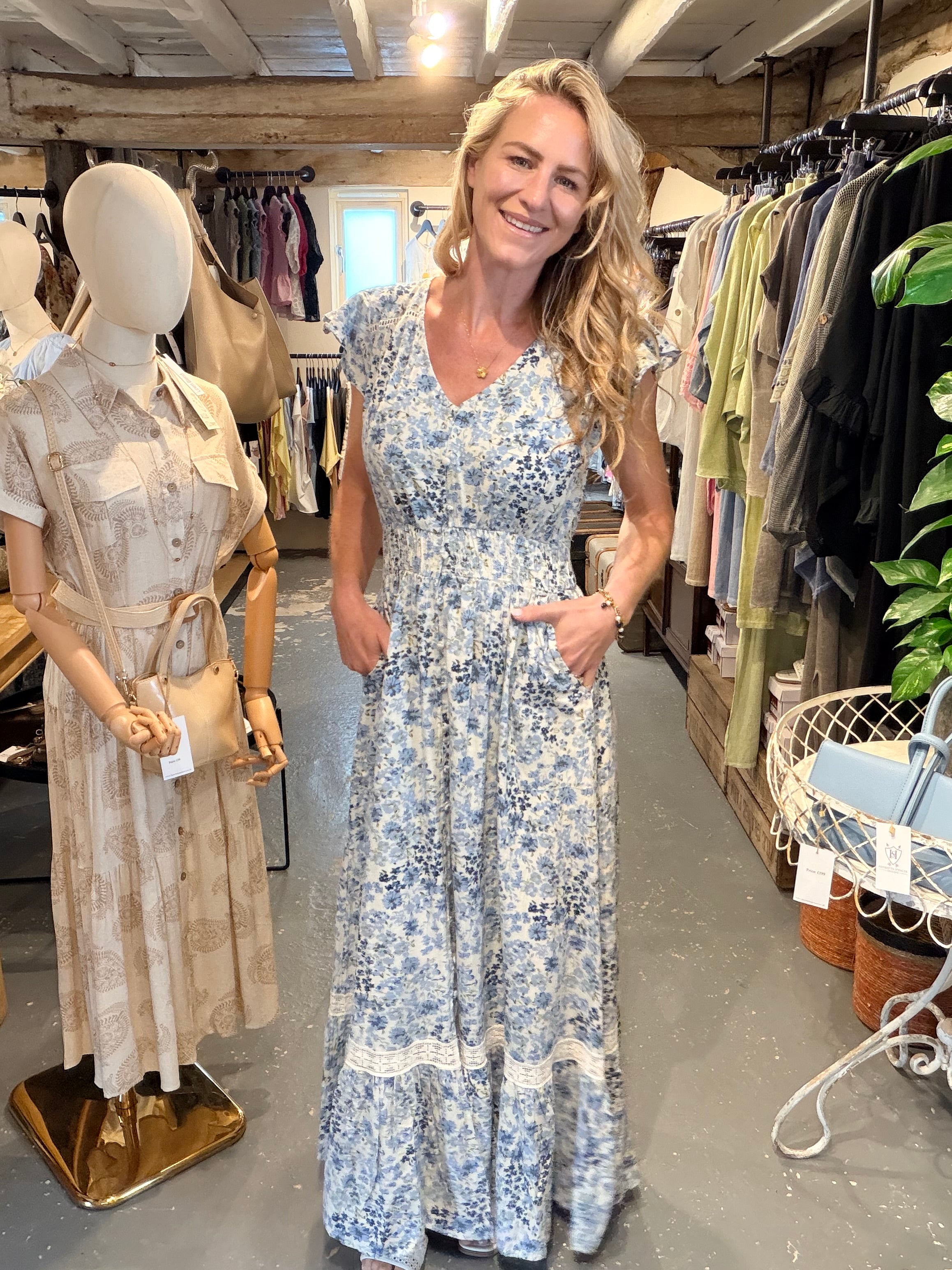 A woman stands in a clothing boutique wearing a blue and white floral print maxi dress with pockets. A mannequin on the left displays a beige patterned shirt dress, and racks of clothes are visible in the background.