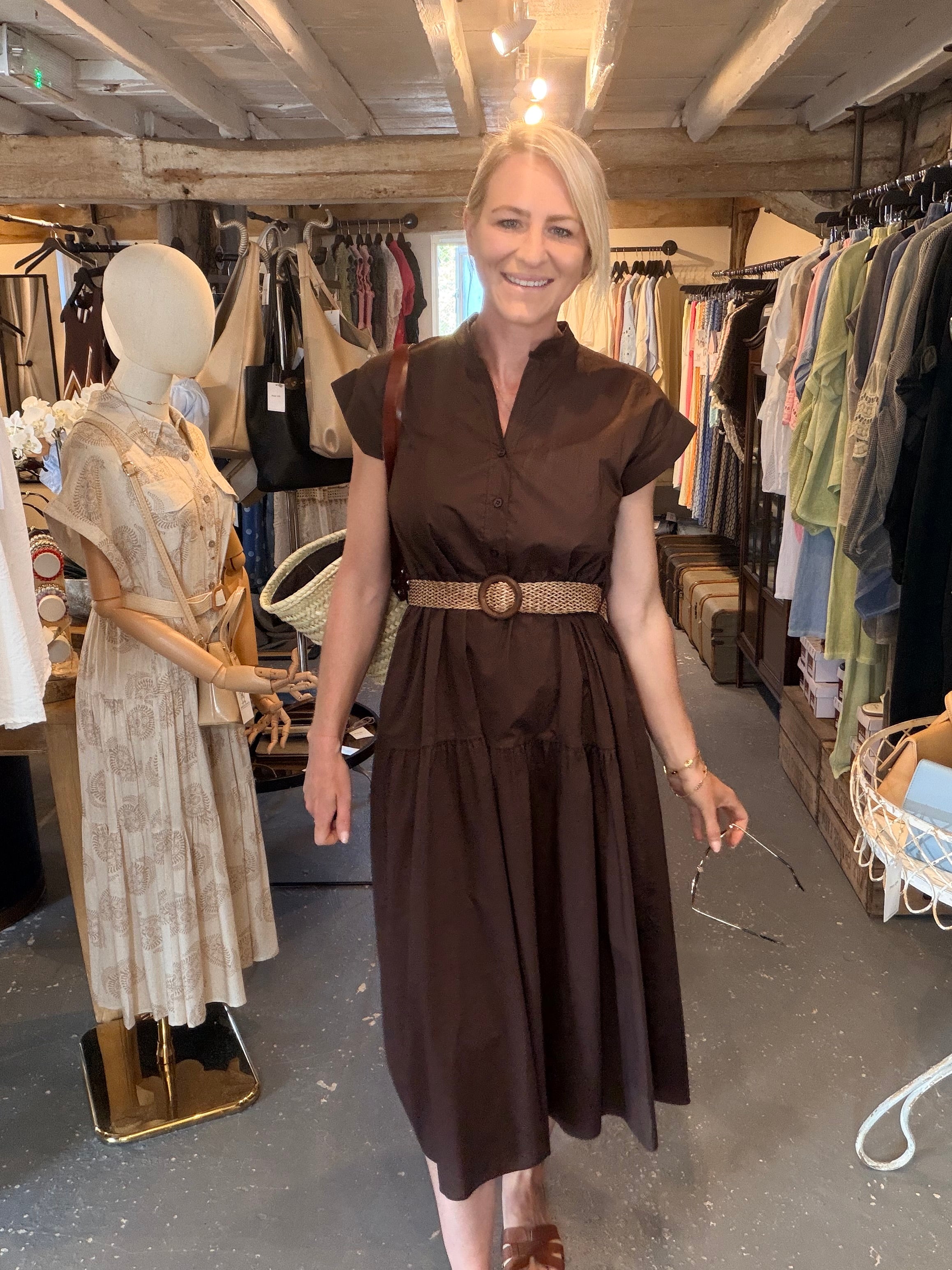 A woman wearing a dark brown midi dress with a textured woven belt stands inside a clothing boutique. The dress has a v-neckline, short cap sleeves, and a tiered skirt.