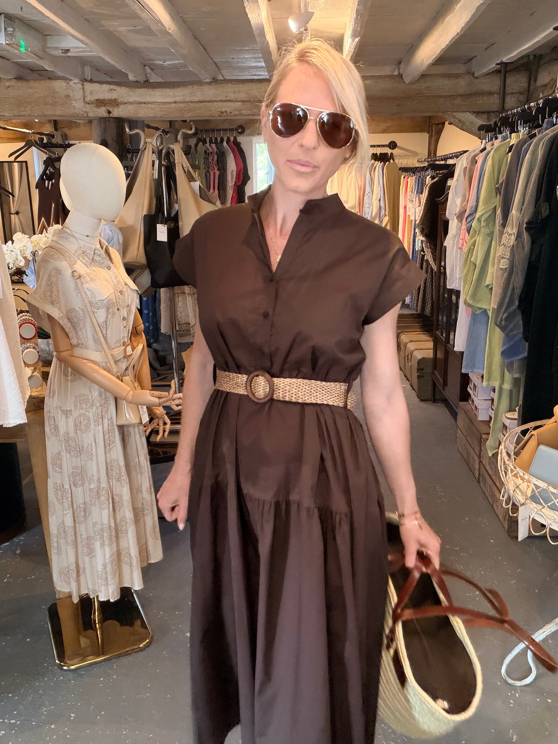 A woman wearing a dark brown short-sleeve midi dress with a textured woven belt stands inside a clothing boutique. She is holding a woven straw tote bag with brown handles.