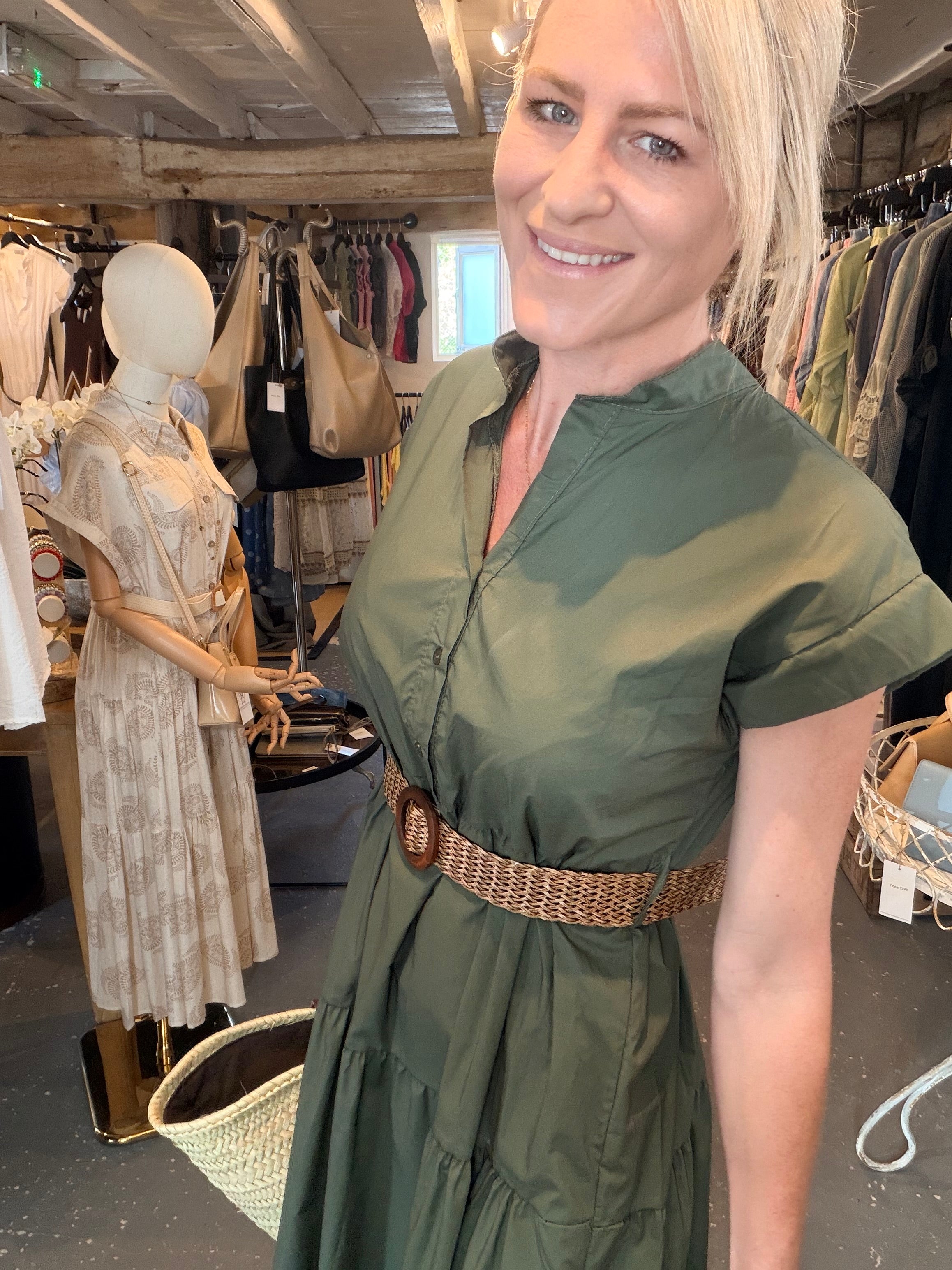 A woman with blonde hair wearing an olive green short-sleeve dress with a brown woven belt, standing inside a clothing boutique.