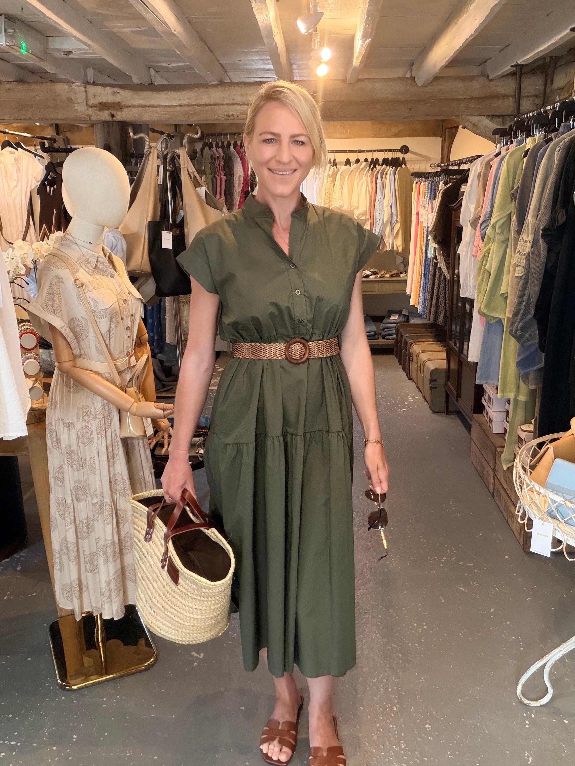 A woman stands in a clothing boutique wearing an olive green midi dress with a brown belt, holding a straw tote bag. The boutique has clothing racks on both sides and a mannequin dressed in a light patterned dress to her left.