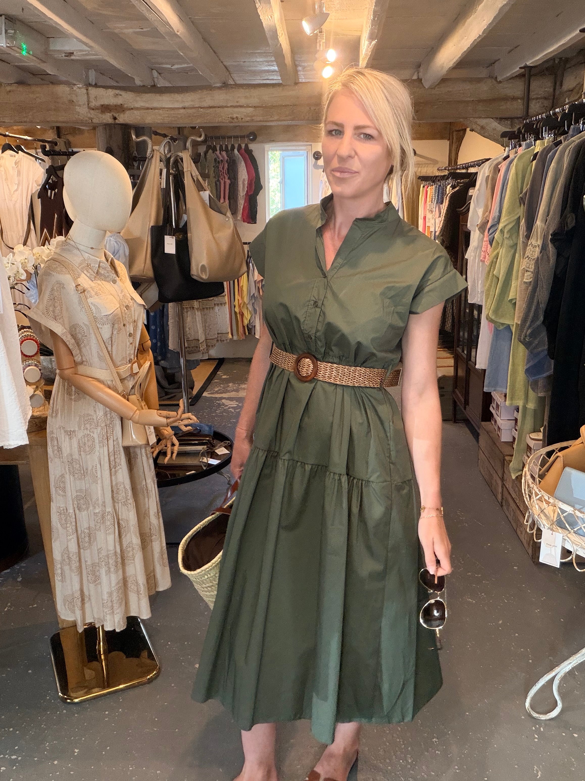 A woman wearing an olive green midi dress with a brown belt stands inside a clothing boutique. The dress has short sleeves, a shawl collar, and a tiered skirt.
