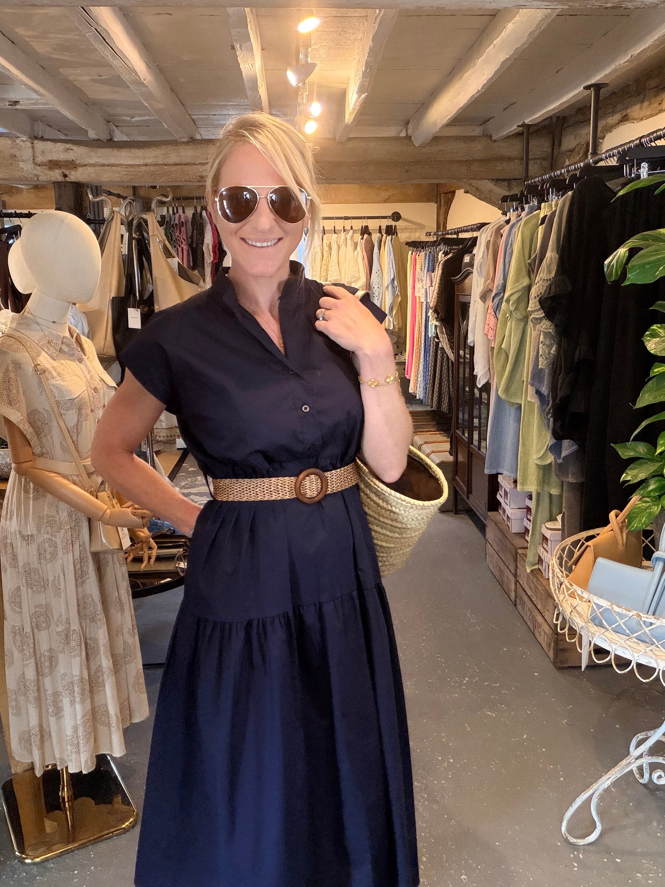 A woman wearing a navy blue tiered midi dress with a tan woven belt and a large round buckle, standing inside a clothing boutique. She is wearing round sunglasses and holding a woven straw bag.