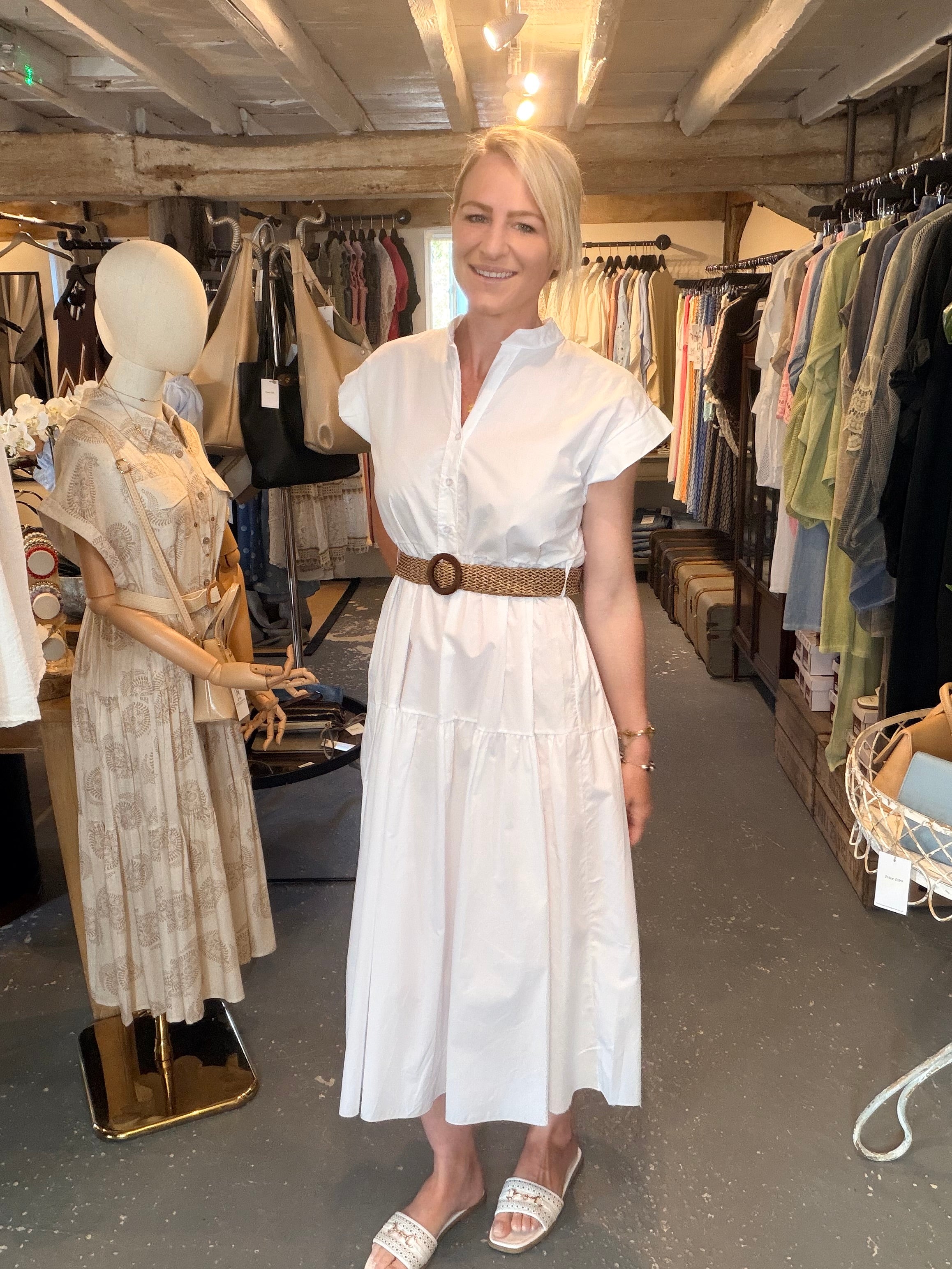 A woman with blonde hair stands in a clothing boutique wearing a white short-sleeve midi dress with a brown woven belt and white slide sandals. A mannequin in a beige patterned dress stands to her left, with racks of clothes in the background.