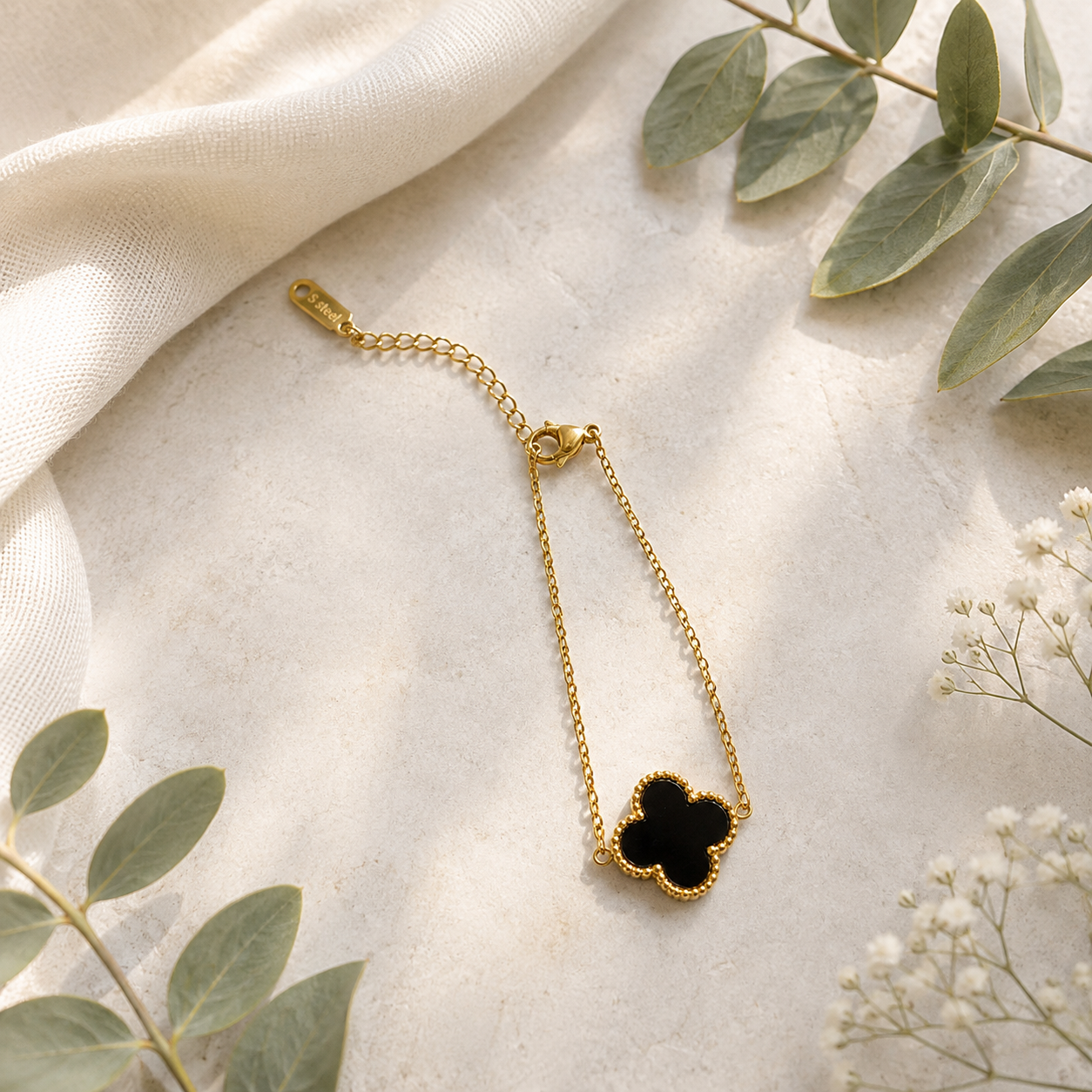 A delicate gold chain bracelet with a black clover-shaped pendant, styled on a textured cream surface with soft fabric and green leaves.