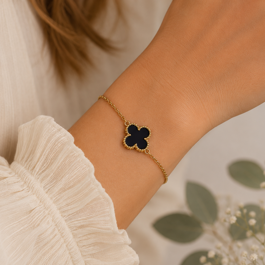 A delicate gold chain bracelet with a black clover charm, worn on a woman's wrist. The image features a close-up of the wrist and a cream-colored ruffled blouse, with soft-focus green leaves in the background.