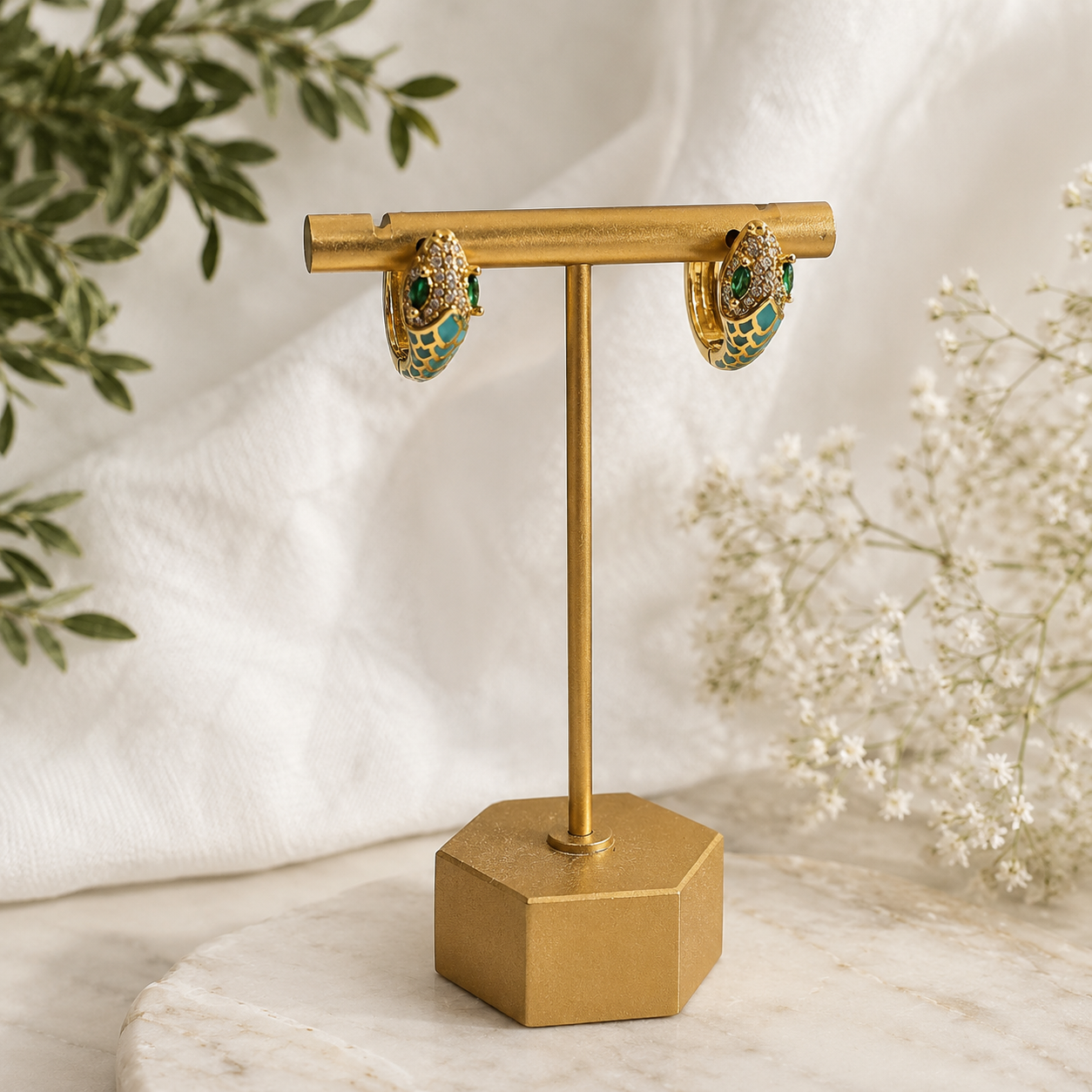 A pair of gold hoop earrings with green and white enamel details, displayed on a gold stand against a soft white fabric backdrop with decorative greenery and baby's breath flowers.