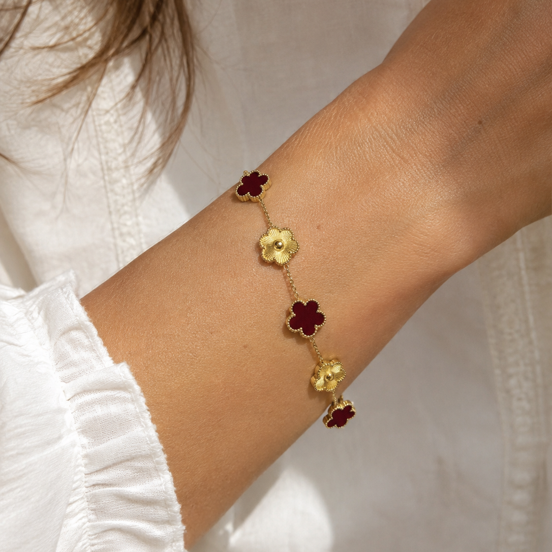 A gold chain bracelet with alternating red and gold four-petal flower charms, worn on a person's wrist over a white ruffled blouse.