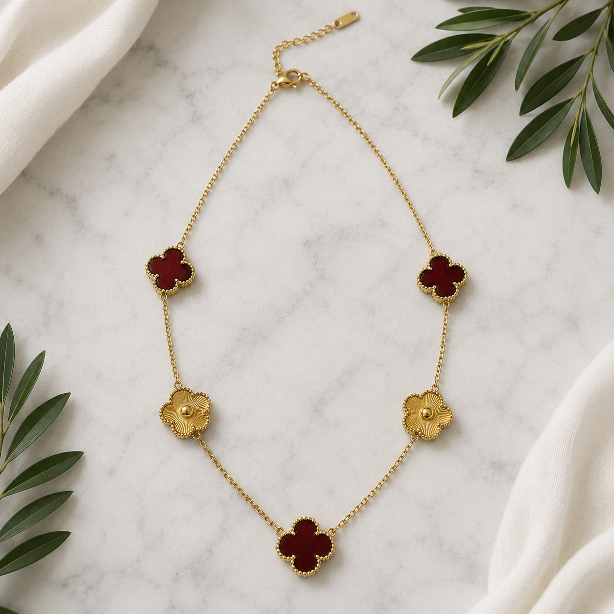 A gold chain necklace with five clover-shaped charms, featuring deep red enamel centers and textured gold edges, styled on a white marble surface with white fabric and green leaves.