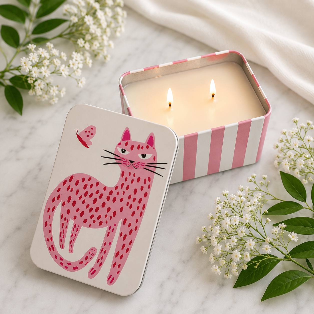 A pink and white striped tin candle with two lit wicks sits on a marble surface next to its illustrated lid, which features a pink spotted cat and butterfly design. White flowers and a white fabric are styled around it.