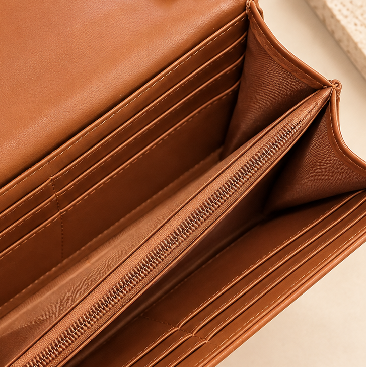 Open tan leather wallet with multiple card slots, a zip compartment, and gold-tone hardware, photographed on a light neutral surface with small dried floral props.