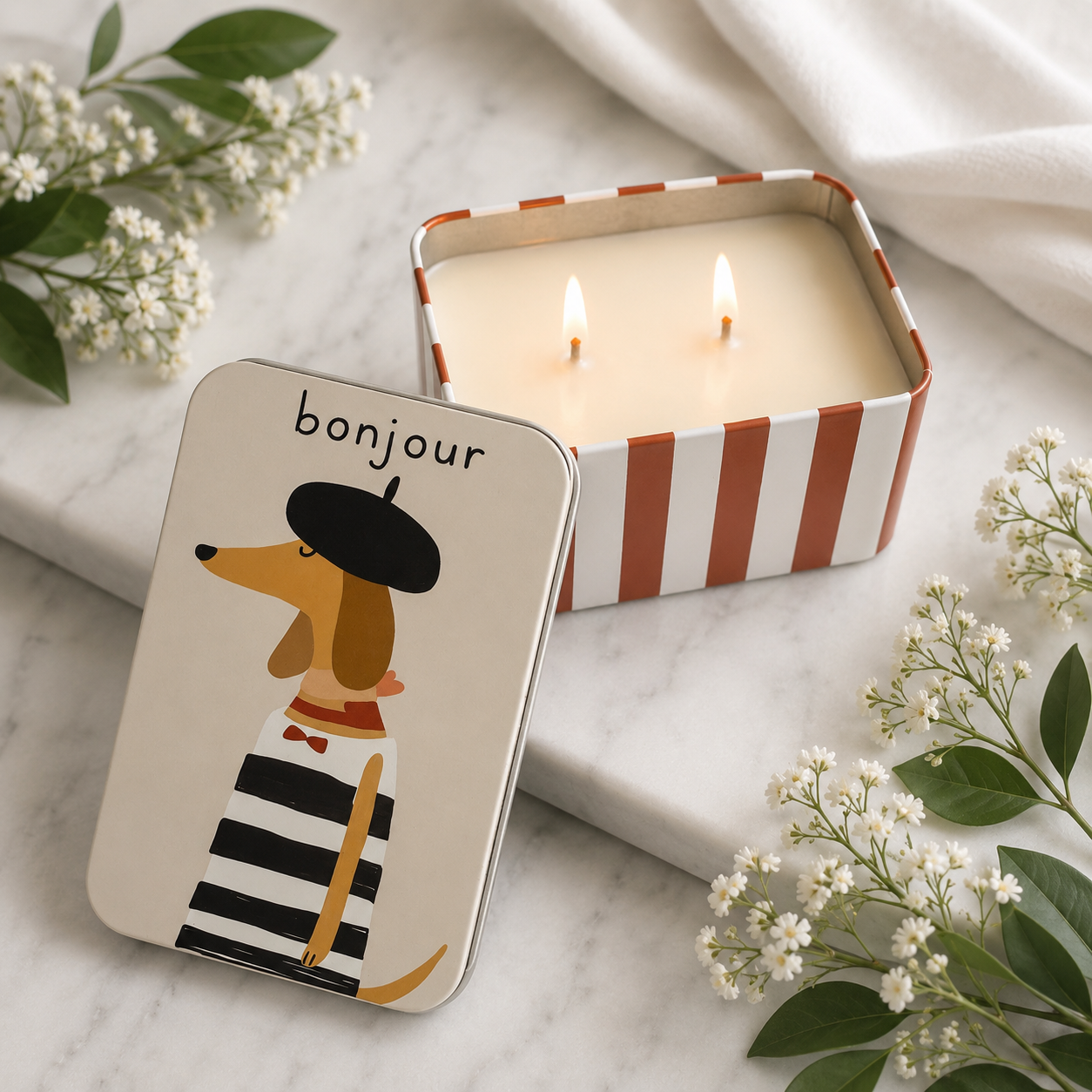 A decorative tin candle shown open with two lit wicks, alongside its illustrated lid. The lid features a dachshund wearing a black beret and black-and-white striped outfit with the word "bonjour" above.