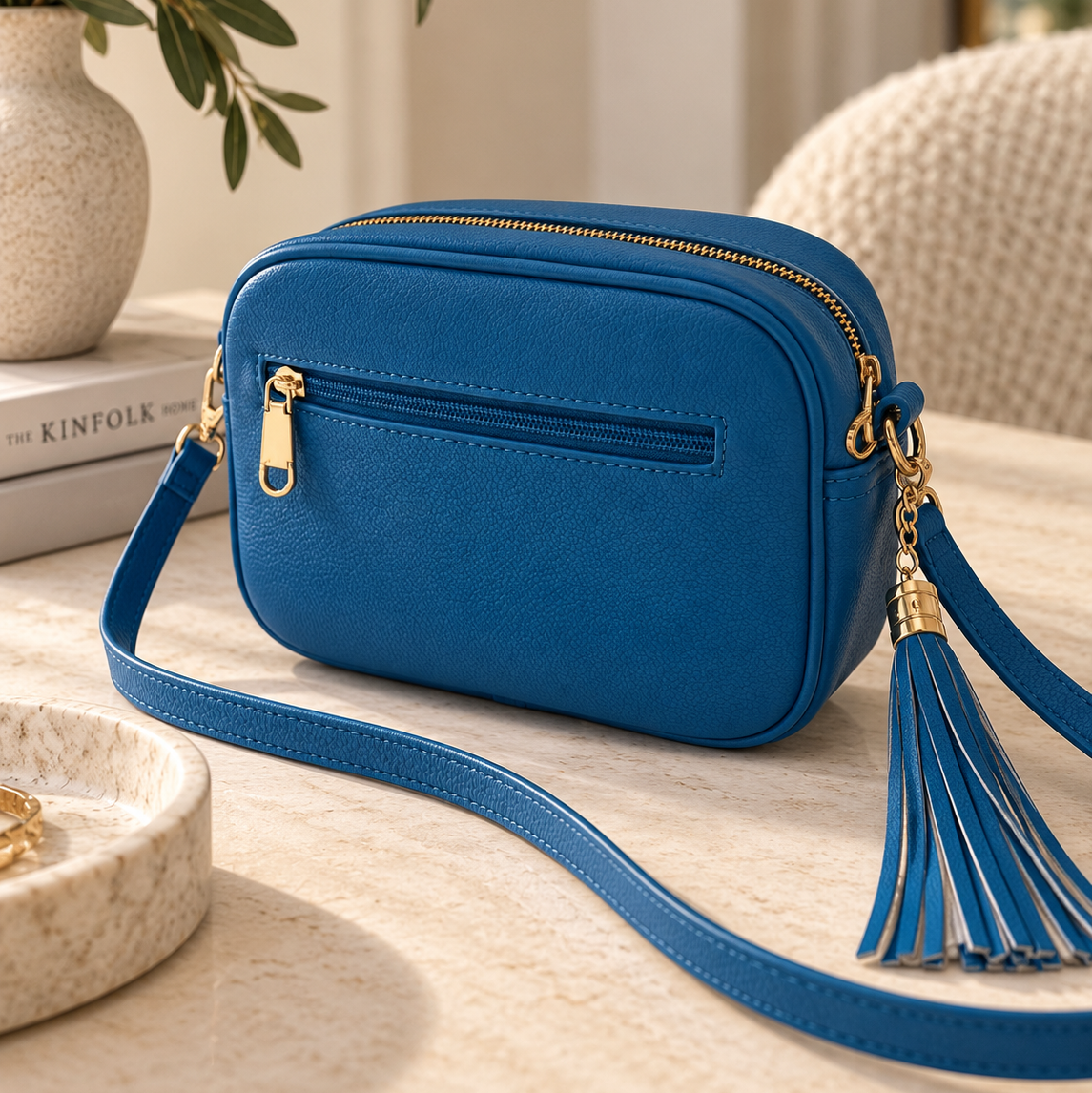 A blue leather crossbody camera bag with gold-tone hardware, a front zip pocket, and a tassel detail, styled on a light stone tabletop with a vase and books in the background.