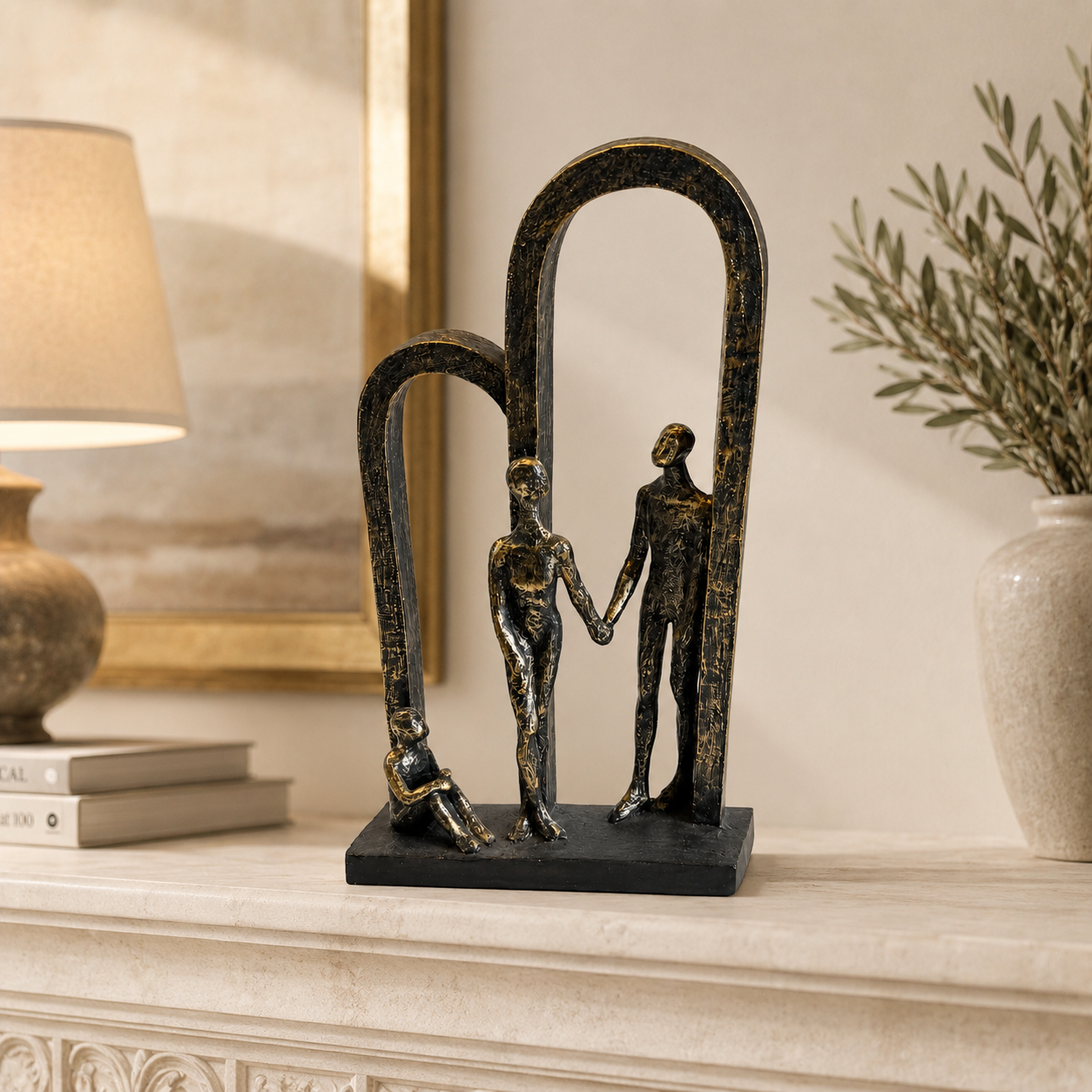 A decorative bronze-finished sculpture of three stylized human figures under two arches, displayed on a light stone mantelpiece in a warmly lit interior setting with a lamp and vase in the background.