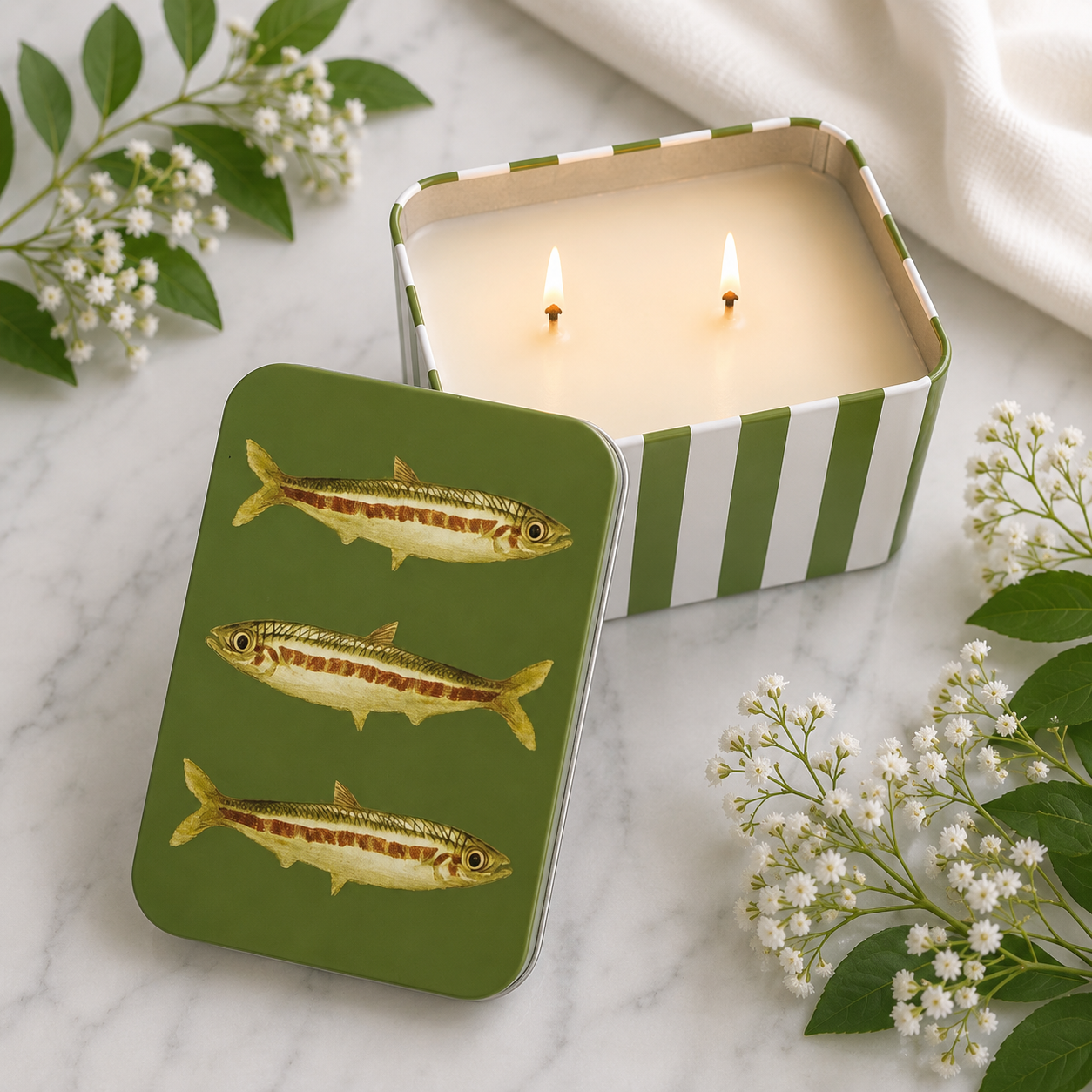 A square tin candle with a green lid decorated with three gold fish illustrations, shown next to an open candle with white wax and two lit wicks. The tins have green and white stripes, set on a white marble surface with baby's breath flowers and a white fabric background.