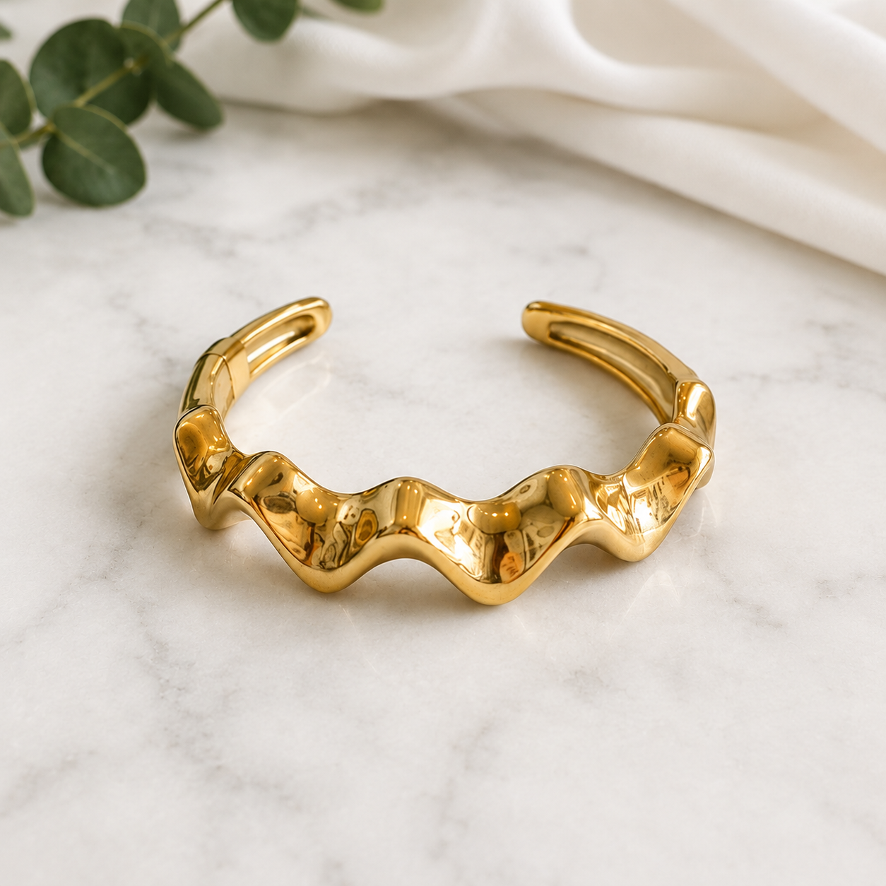 A polished gold cuff bracelet with a wavy, organic textured design, displayed on a white marble surface with a white fabric and green eucalyptus leaves in the background.