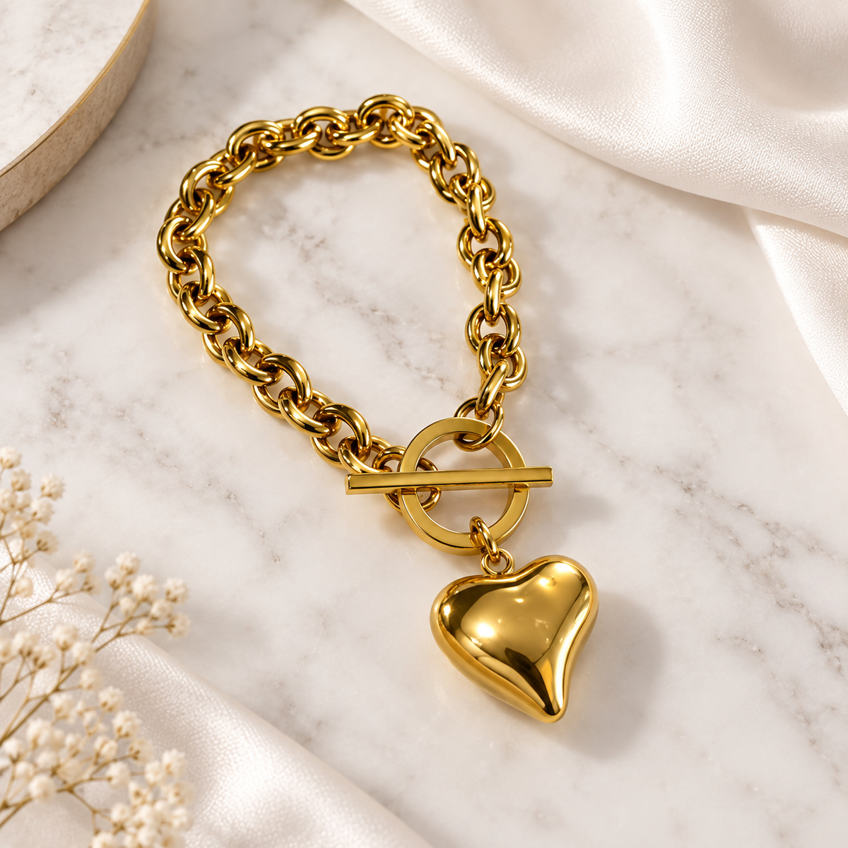 A gold-toned chain bracelet with a toggle clasp and a dangling heart-shaped charm, styled on a white marble surface with satin fabric and small white flowers.