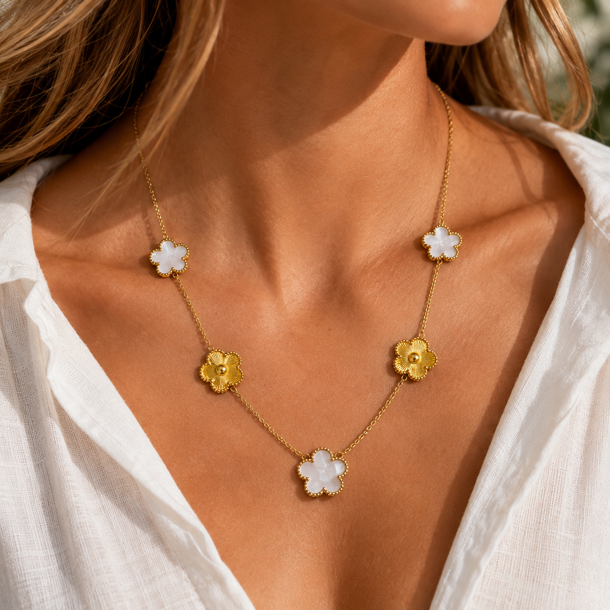 A close-up of a woman wearing a delicate gold chain necklace with five flower-shaped charms, three in white and two in gold, styled with a white blouse.