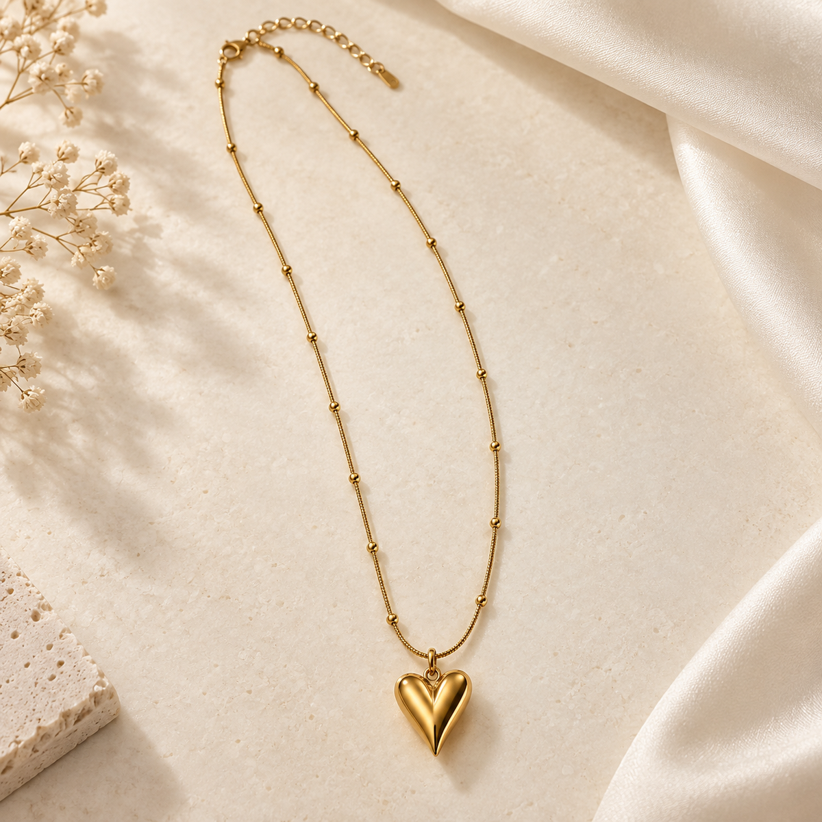 A gold necklace with a textured, beaded chain and a polished heart-shaped pendant, styled on a cream-colored textured surface with soft white fabric and dried baby's breath flowers.