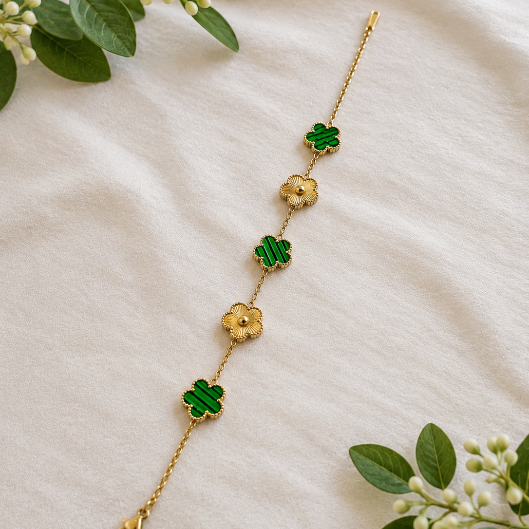 A delicate gold chain bracelet featuring four green enamel clover motifs and three gold flower charms, laid out on a soft beige fabric background with decorative green leaves.
