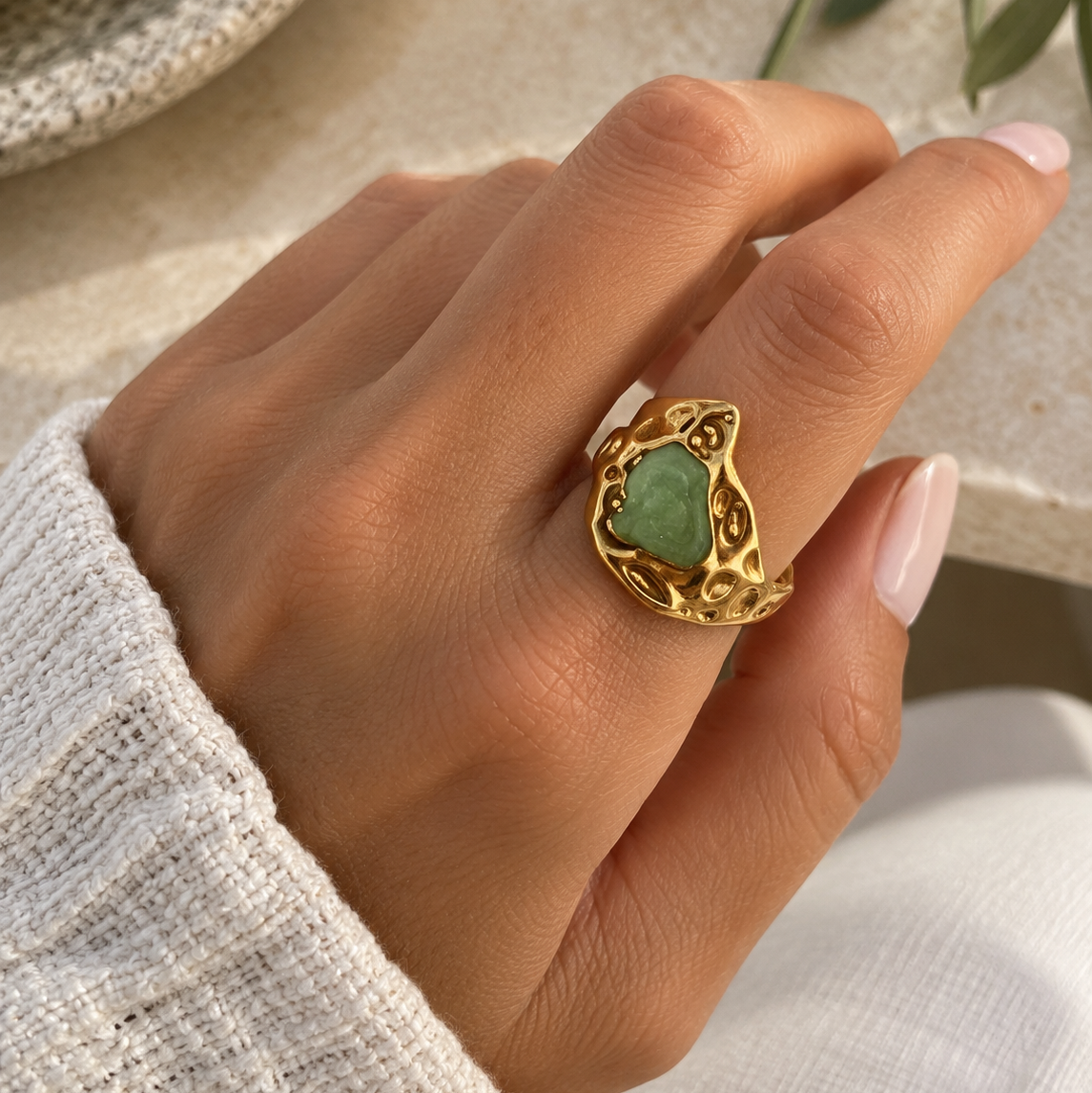 A gold ring with a heart-shaped green stone, worn on a hand with light pink manicured nails and a white knit sweater sleeve.