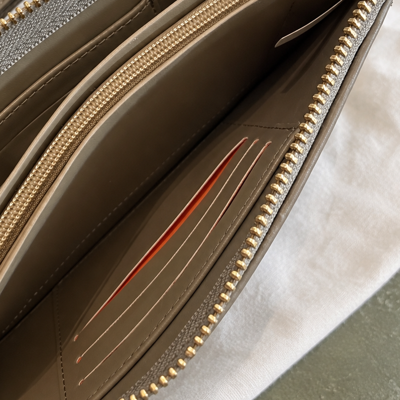 An open taupe leather zip-around wallet with gold-tone hardware, displayed on a white fabric background with a leafy sprig in the corner.
