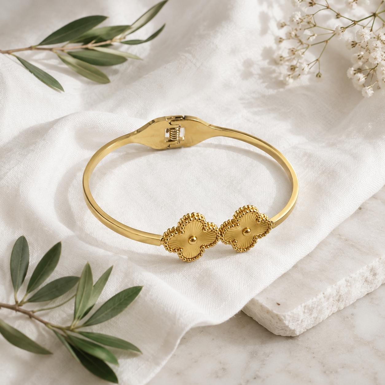 A gold bangle bracelet with two textured clover motifs, displayed on a white linen cloth with olive branches and baby's breath flowers.