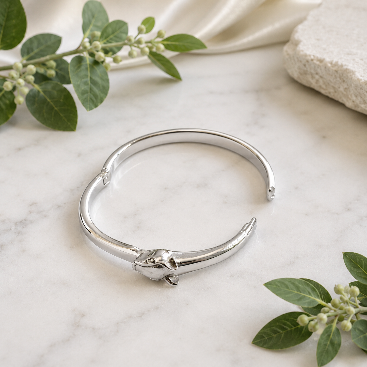 A polished silver-toned open cuff bangle bracelet with a sculpted animal head at the hinge, displayed on a white marble surface with green leaves and a stone prop.