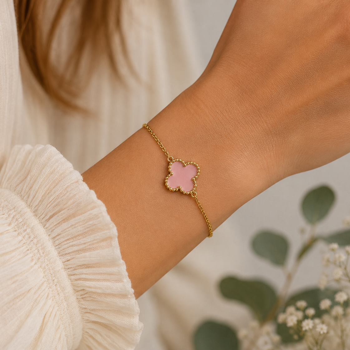 A gold chain bracelet with a pink clover-shaped charm, worn on a woman's wrist. The image features a close-up of the wrist and hand, with a soft-focus background of white flowers and green leaves.
