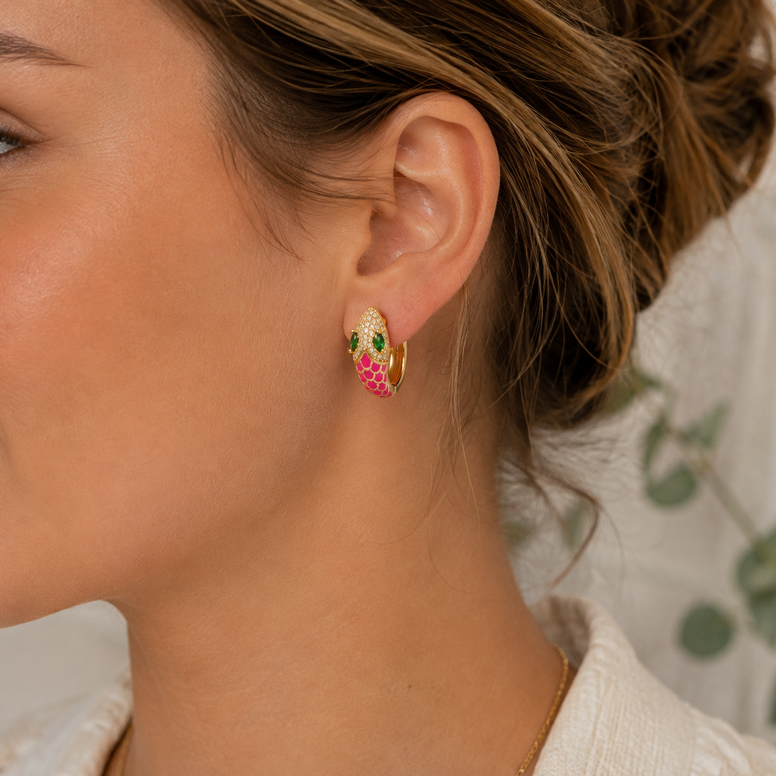 A close-up of a woman's ear wearing a small gold huggie hoop earring adorned with pink and green enamel details.